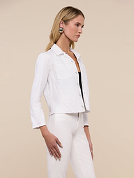Wearing the Janelle Jean Jacket in a feminine fit, a woman with long blonde hair pairs it with white stretch denim pants, a black top, a belt, and statement earrings as she stands against a plain light background.