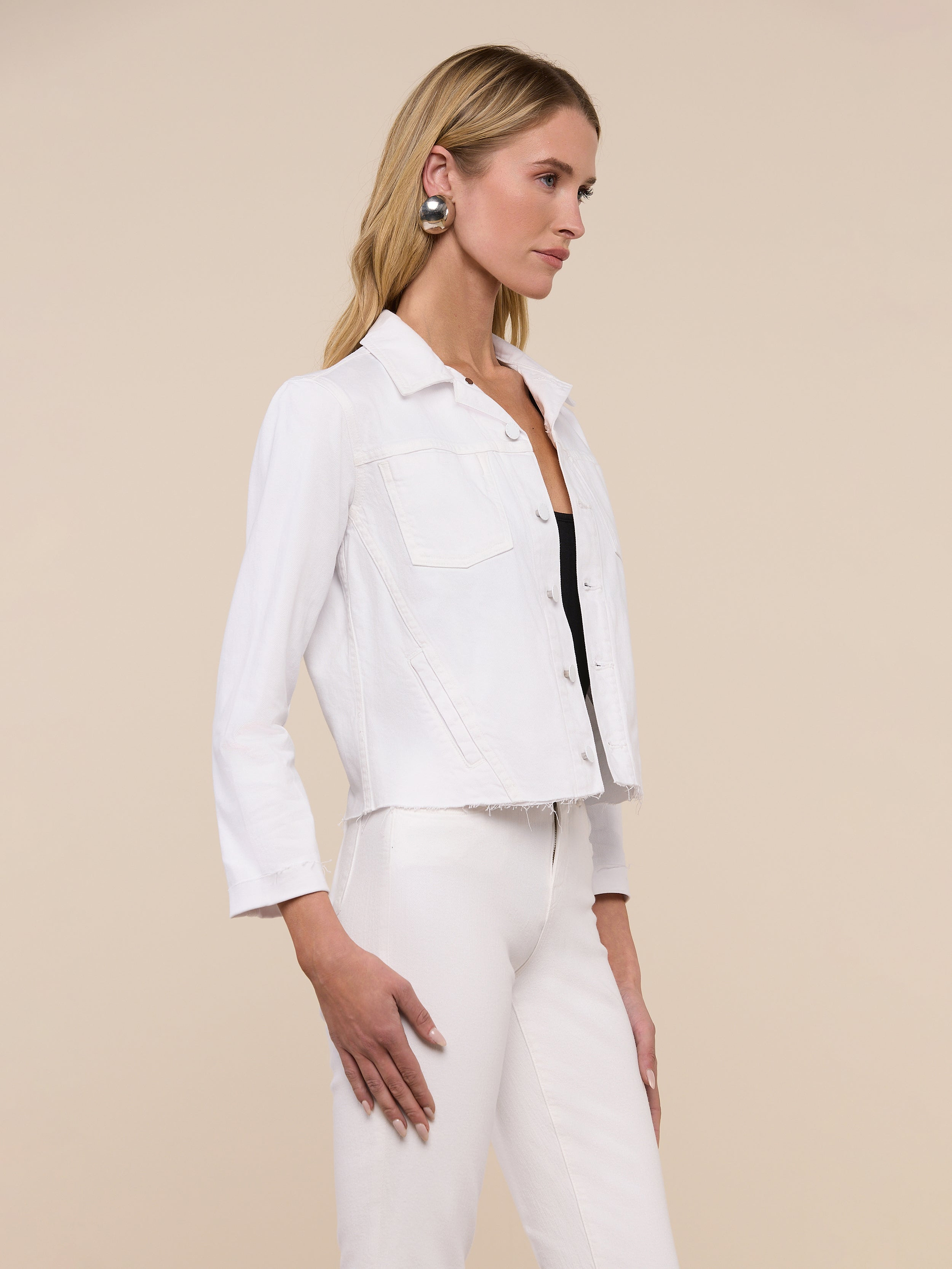 Wearing the Janelle Jean Jacket in a feminine fit, a woman with long blonde hair pairs it with white stretch denim pants, a black top, a belt, and statement earrings as she stands against a plain light background.