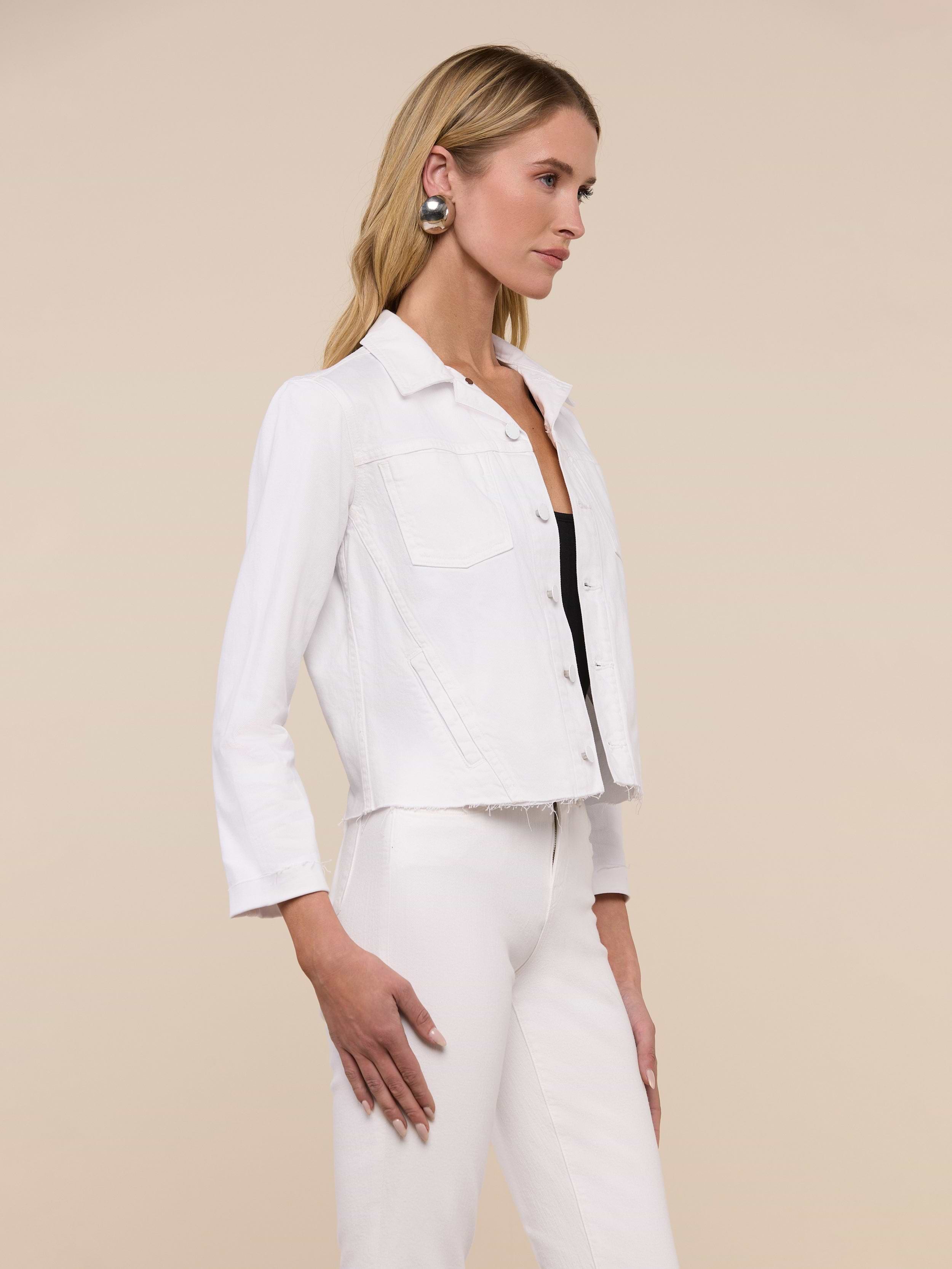 Wearing the Janelle Jean Jacket in a feminine fit, a woman with long blonde hair pairs it with white stretch denim pants, a black top, a belt, and statement earrings as she stands against a plain light background.