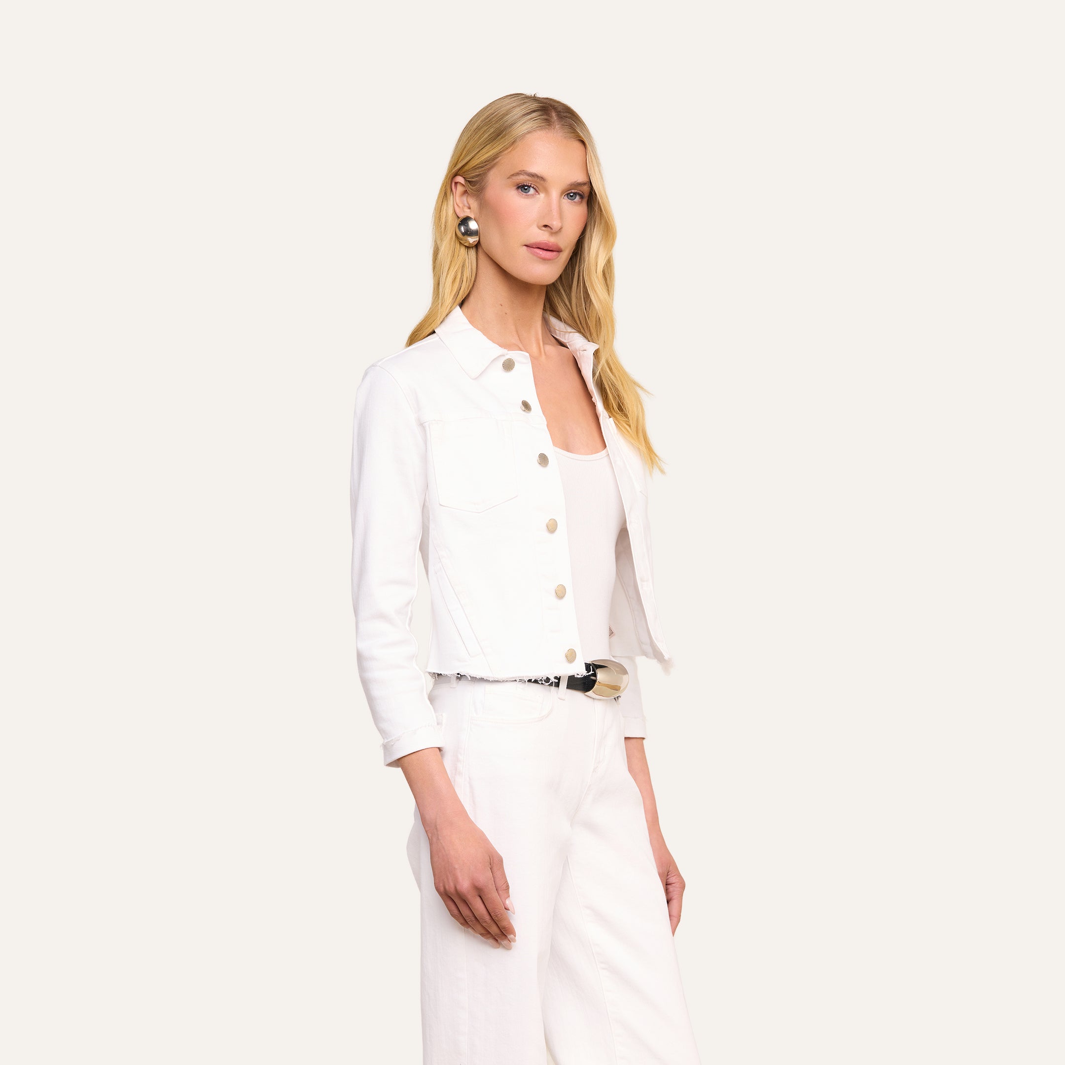 Indossando la giacca Janelle Jean, una donna dai lunghi capelli biondi ne mette in risalto la linea femminile e il denim bianco elasticizzato, abbinata a un top e a pantaloni bianchi coordinati, in piedi su uno sfondo beige.