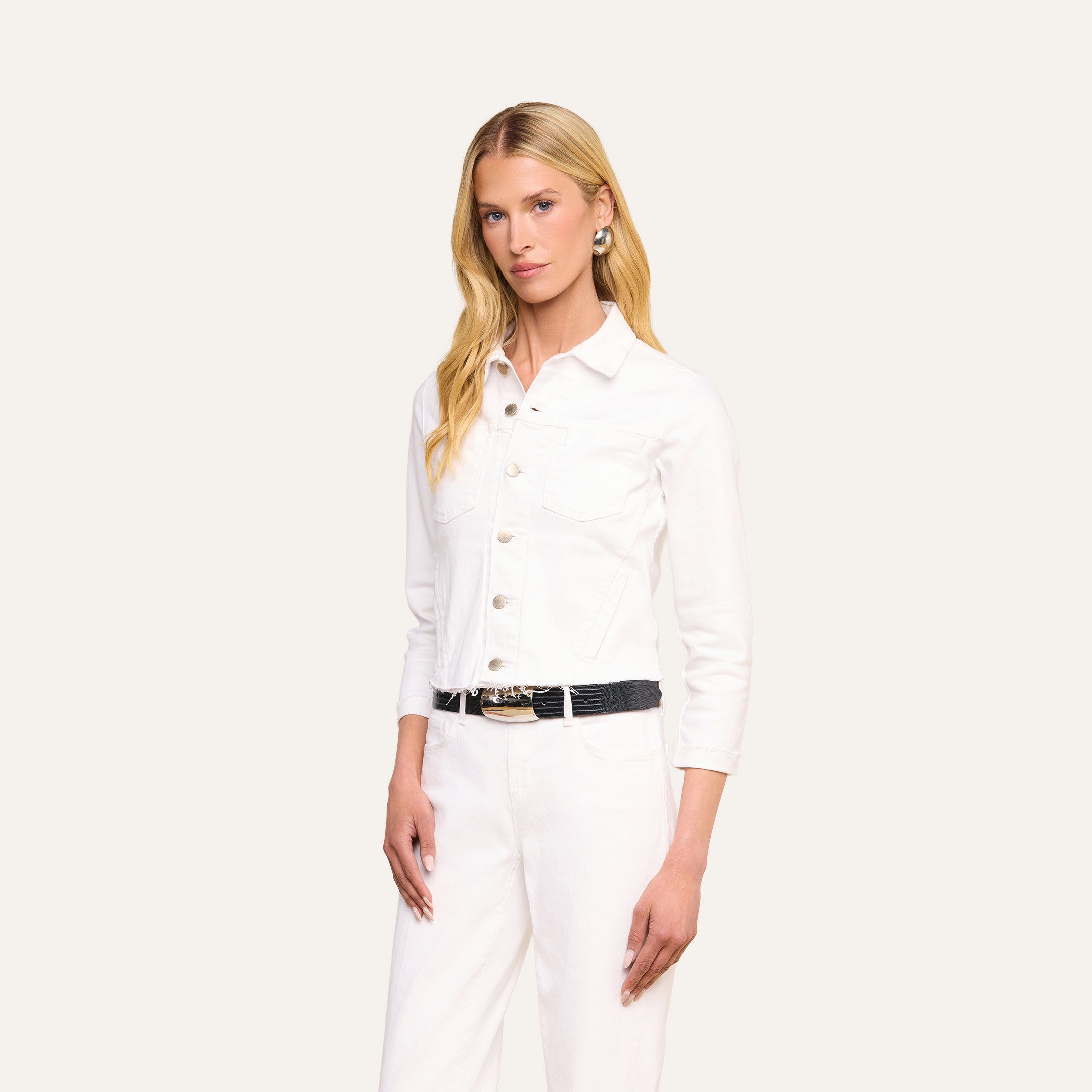 Una donna dai lunghi capelli biondi indossa la giacca Janelle Jean in una versione femminile in denim bianco, abbinata a pantaloni bianchi coordinati e a una cintura nera, mentre posa su uno sfondo beige a tinta unita.
