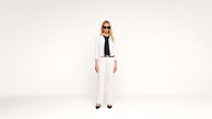 A woman wearing sunglasses, a white jacket, white pants, and black shoes stands against a plain white background.
