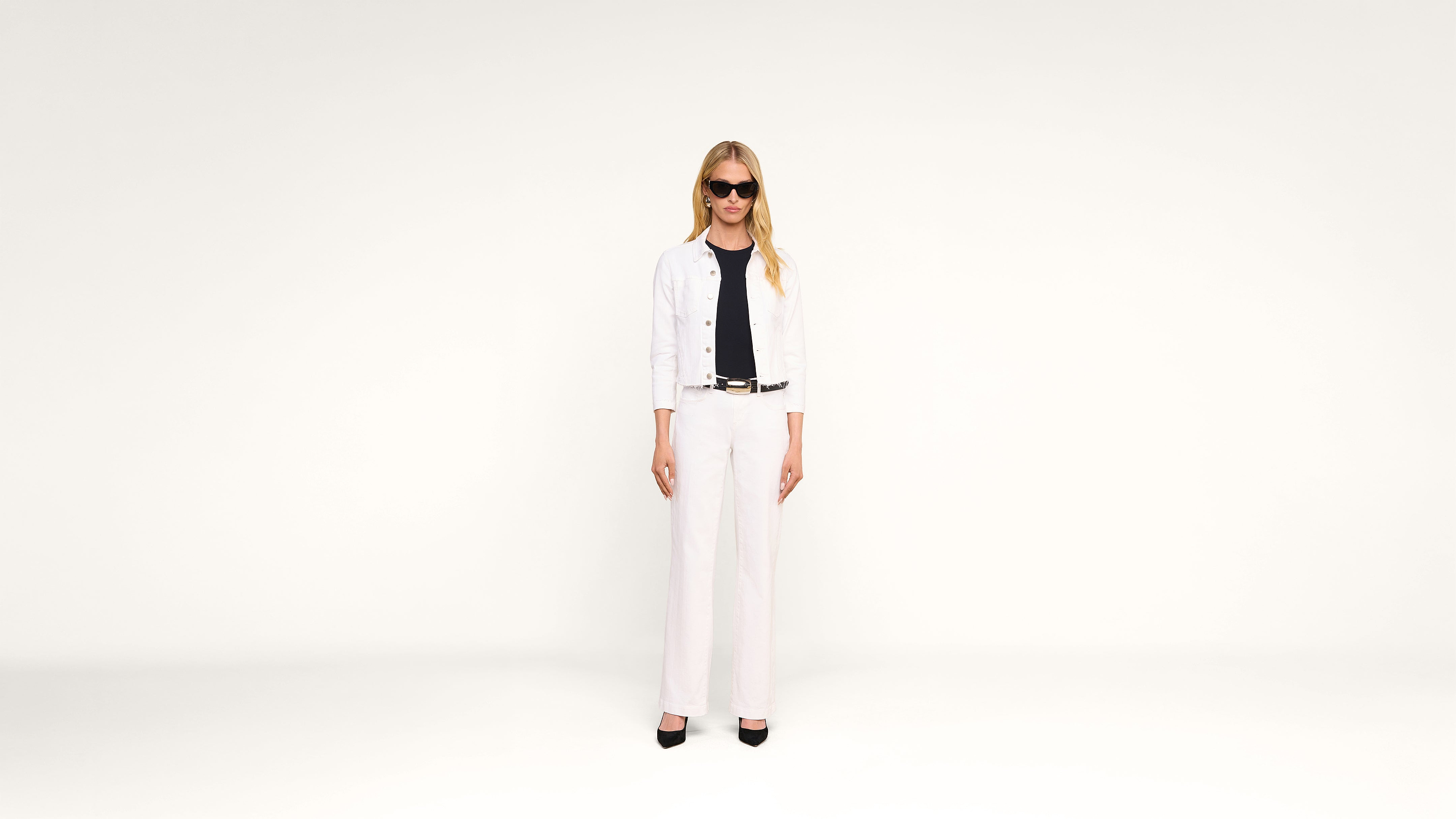 A woman wearing sunglasses, a white jacket, white pants, and black shoes stands against a plain white background.