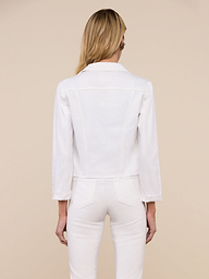 A woman with long blonde hair stands facing away against a plain light background, wearing the Janelle Jean Jacket in stretch white denim with three-quarter sleeves and coordinating white pants. The feminine fit of the jacket enhances her look.