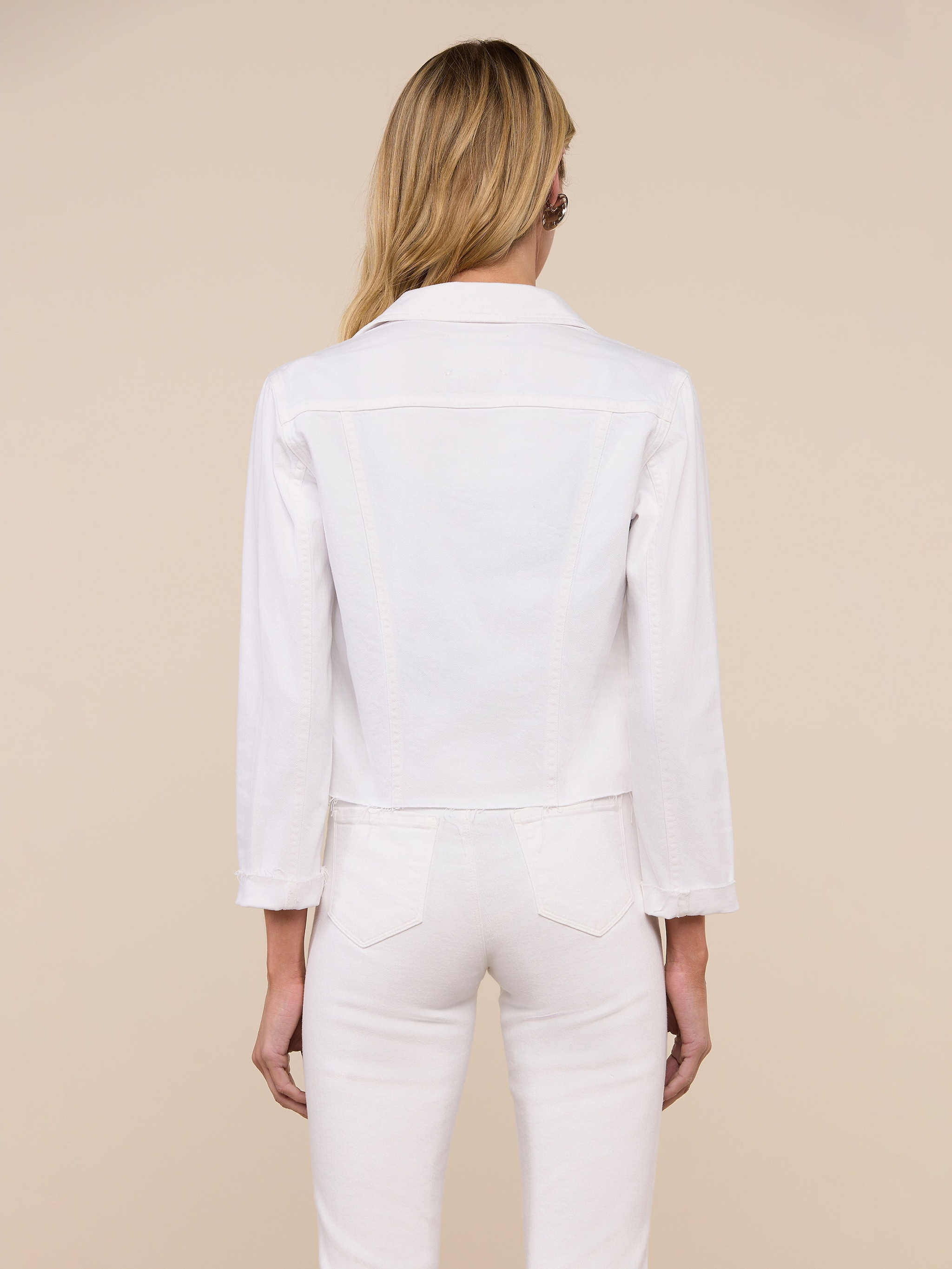 A woman with long blonde hair stands facing away against a plain light background, wearing the Janelle Jean Jacket in stretch white denim with three-quarter sleeves and coordinating white pants. The feminine fit of the jacket enhances her look.