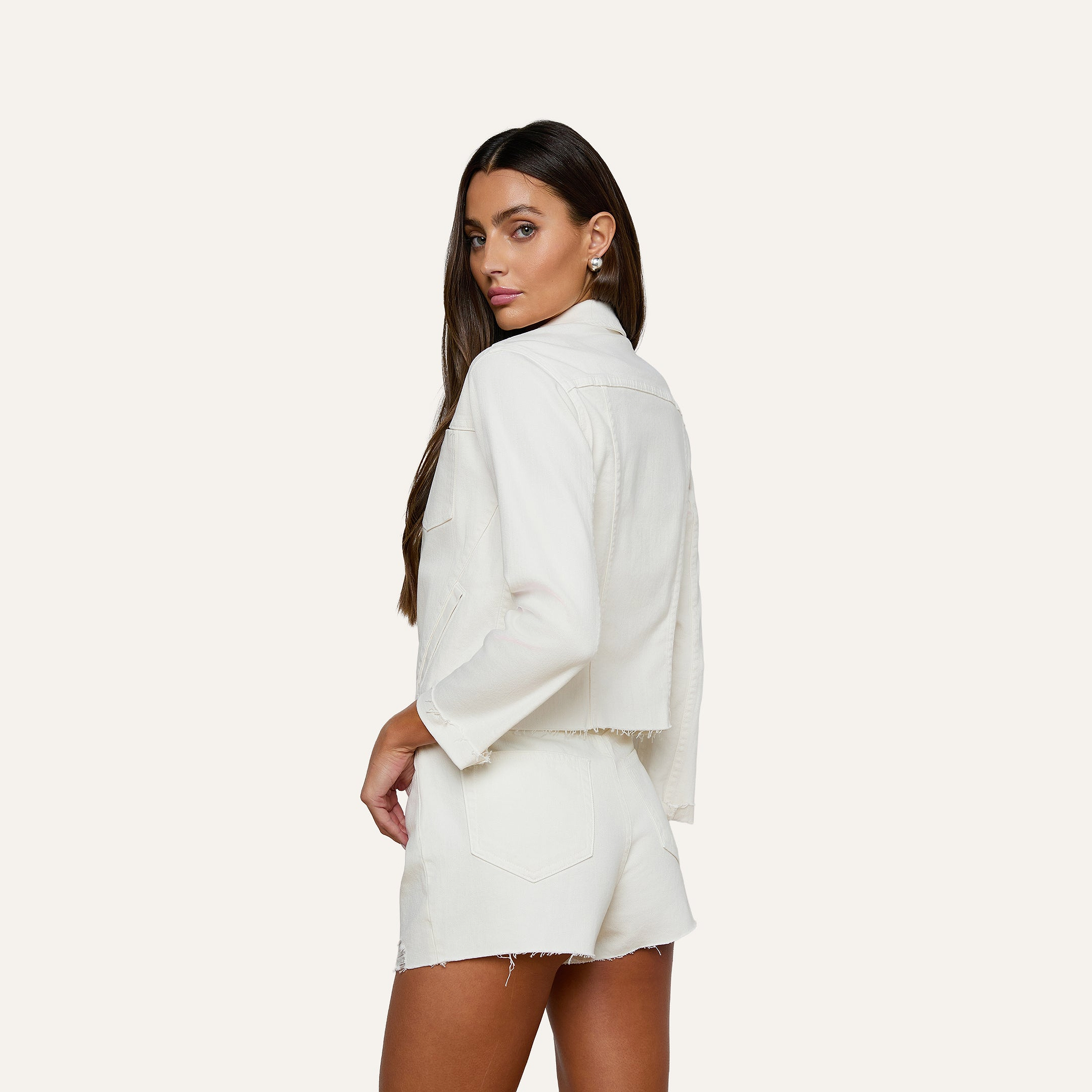 A woman with long brown hair, wearing the slim fit white Janelle Jean Jacket and matching white shorts, stands sideways and looks over her shoulder against a plain white background.