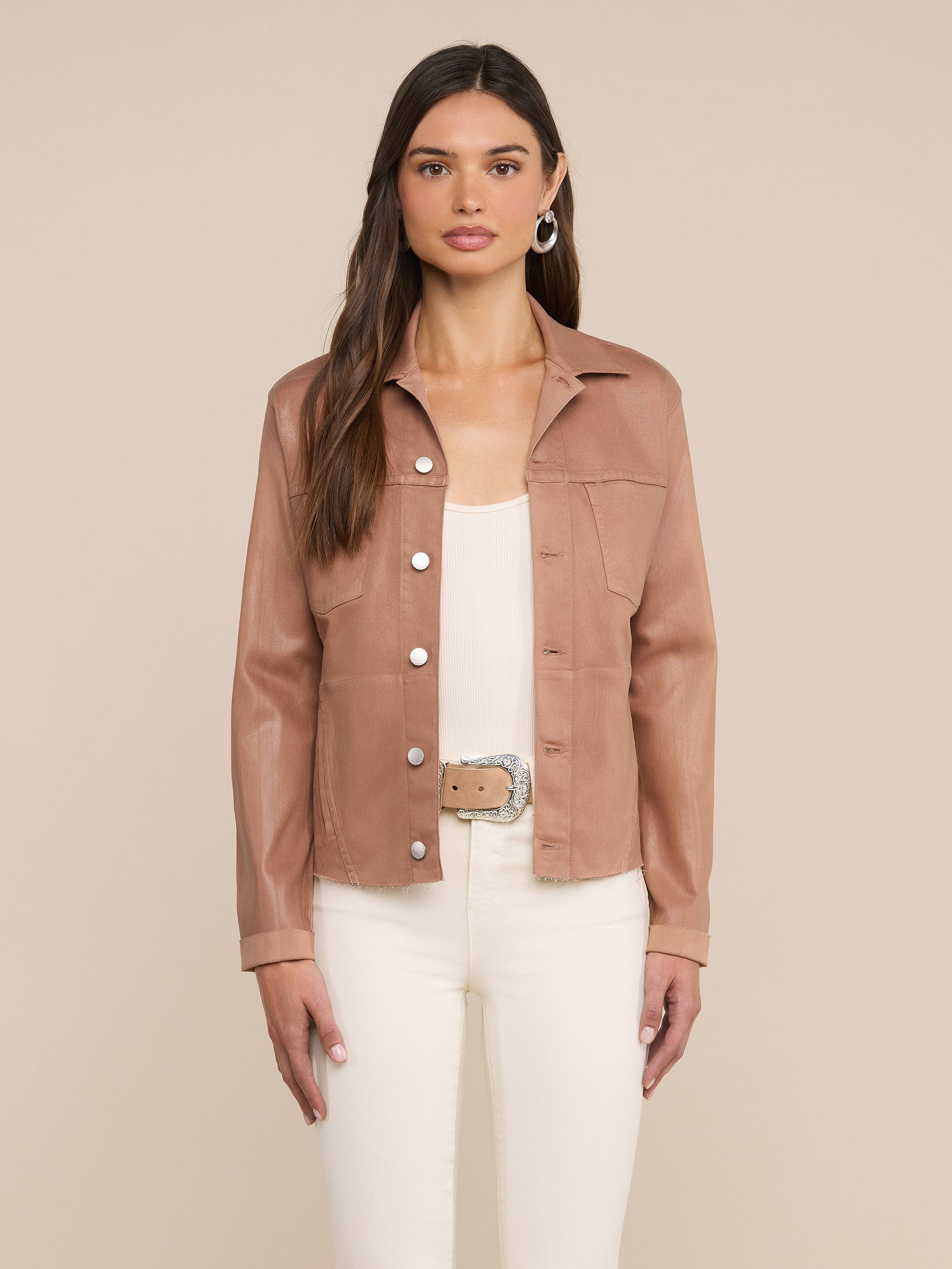 A woman stands facing forward, wearing the Janelle Coated Jean Jacket in hazelnut brown over a white top and white pants with a beige belt, set against a plain light background.