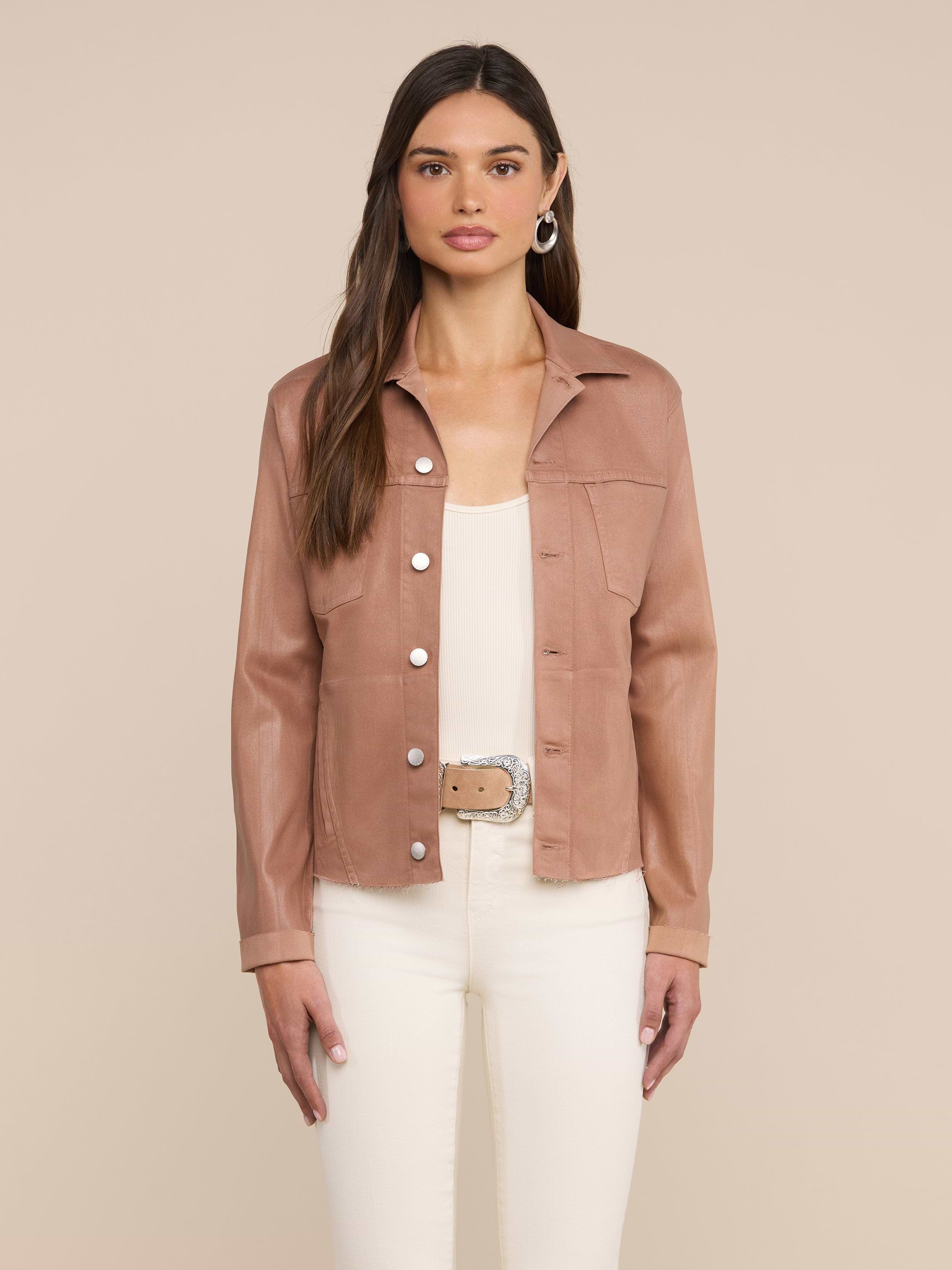 A woman stands facing forward, wearing the Janelle Coated Jean Jacket in hazelnut brown over a white top and white pants with a beige belt, set against a plain light background.