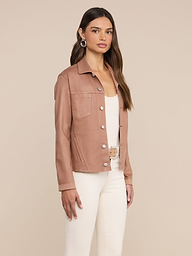 A woman stands against a plain background, wearing the Janelle Coated Jean Jacket in hazelnut brown over a white top and white pants, accessorized with hoop earrings and long straight hair.