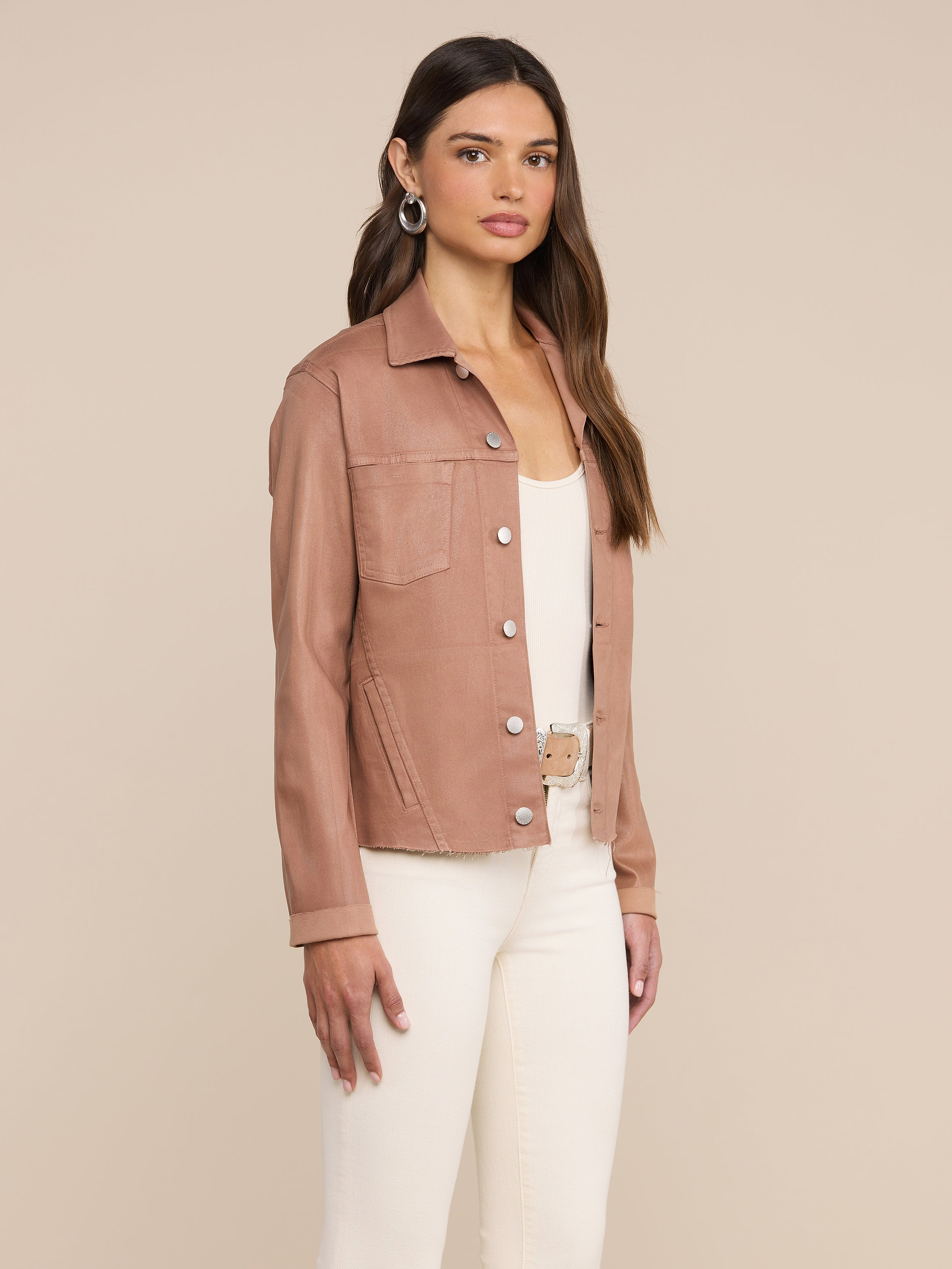 A woman stands against a plain background, wearing the Janelle Coated Jean Jacket in hazelnut brown over a white top and white pants, accessorized with hoop earrings and long straight hair.