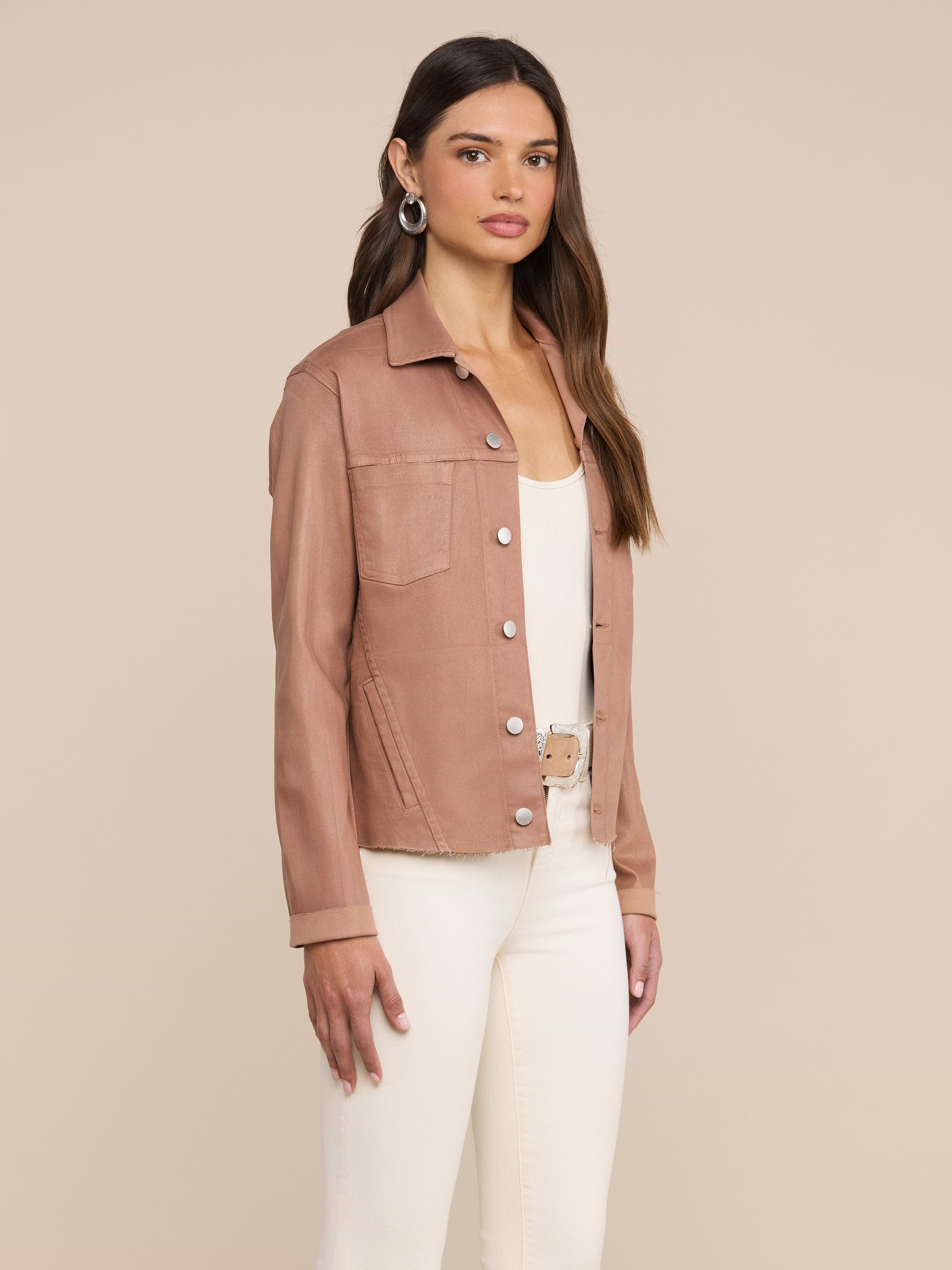 A woman stands against a plain background, wearing the Janelle Coated Jean Jacket in hazelnut brown over a white top and white pants, accessorized with hoop earrings and long straight hair.