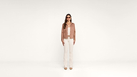 A woman stands against a plain white background, wearing sunglasses, a light brown jacket, a white shirt, white pants, and beige shoes. She faces forward with a neutral expression.