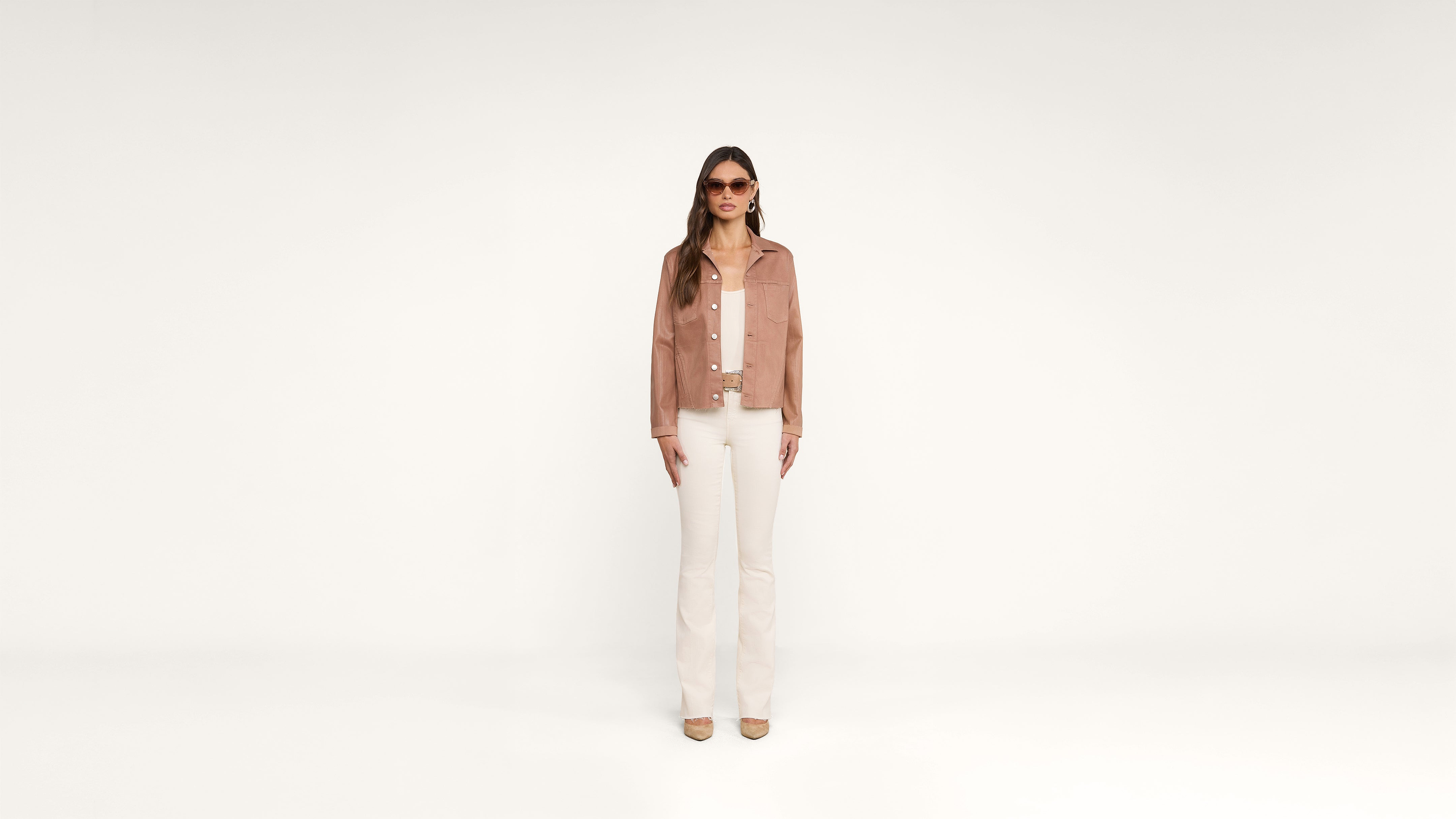 A woman stands against a plain white background, wearing sunglasses, a light brown jacket, a white shirt, white pants, and beige shoes. She faces forward with a neutral expression.