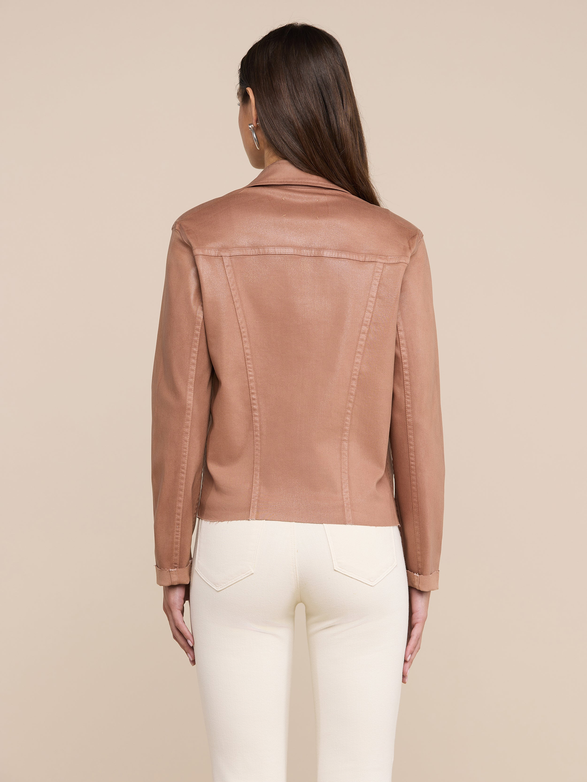 A woman with straight brown hair is shown from the back against a plain, light background, wearing the Janelle Coated Jean Jacket in a slim fit hazelnut brown color paired with white pants.