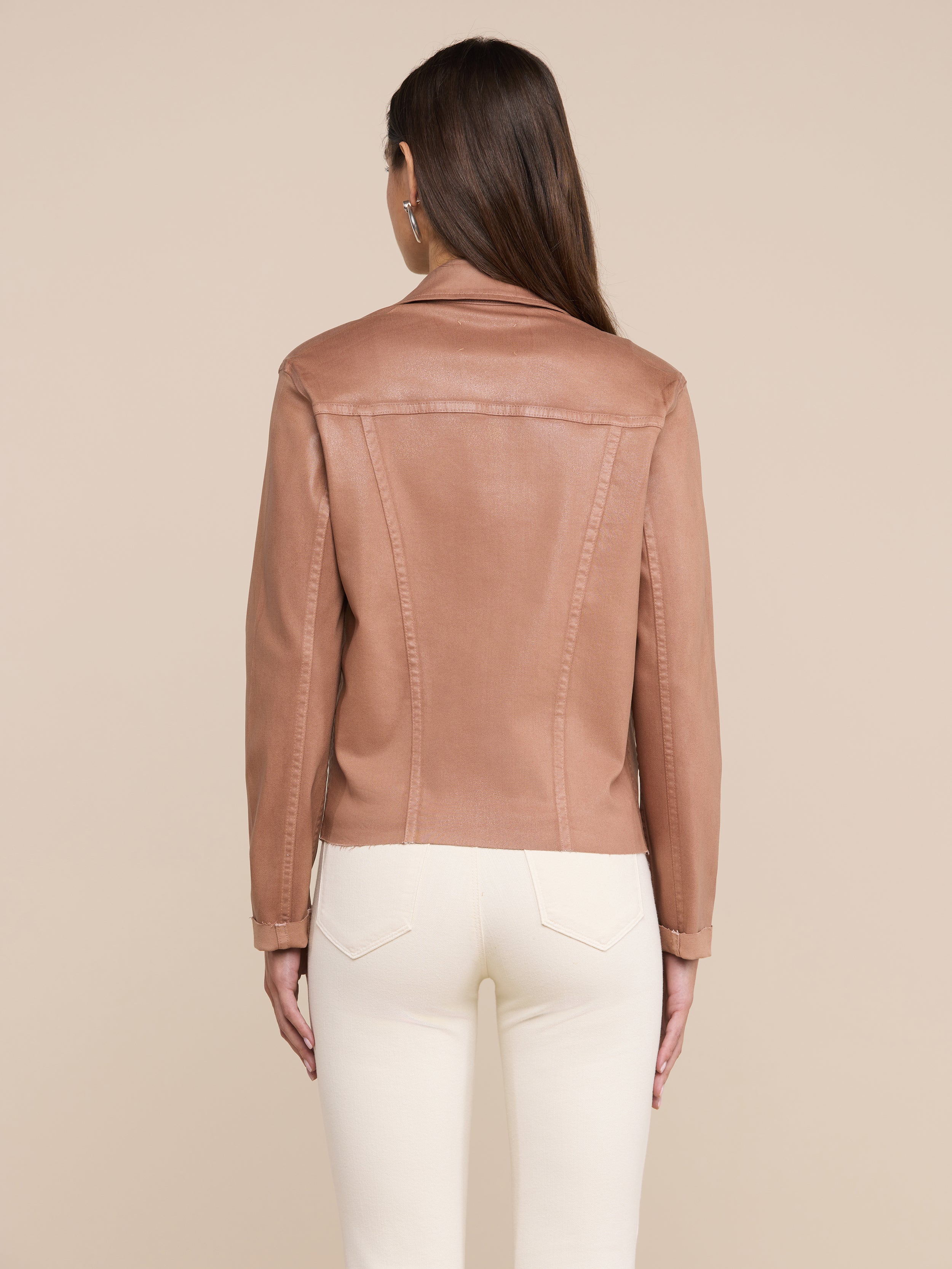 A woman with straight brown hair is shown from the back against a plain, light background, wearing the Janelle Coated Jean Jacket in a slim fit hazelnut brown color paired with white pants.