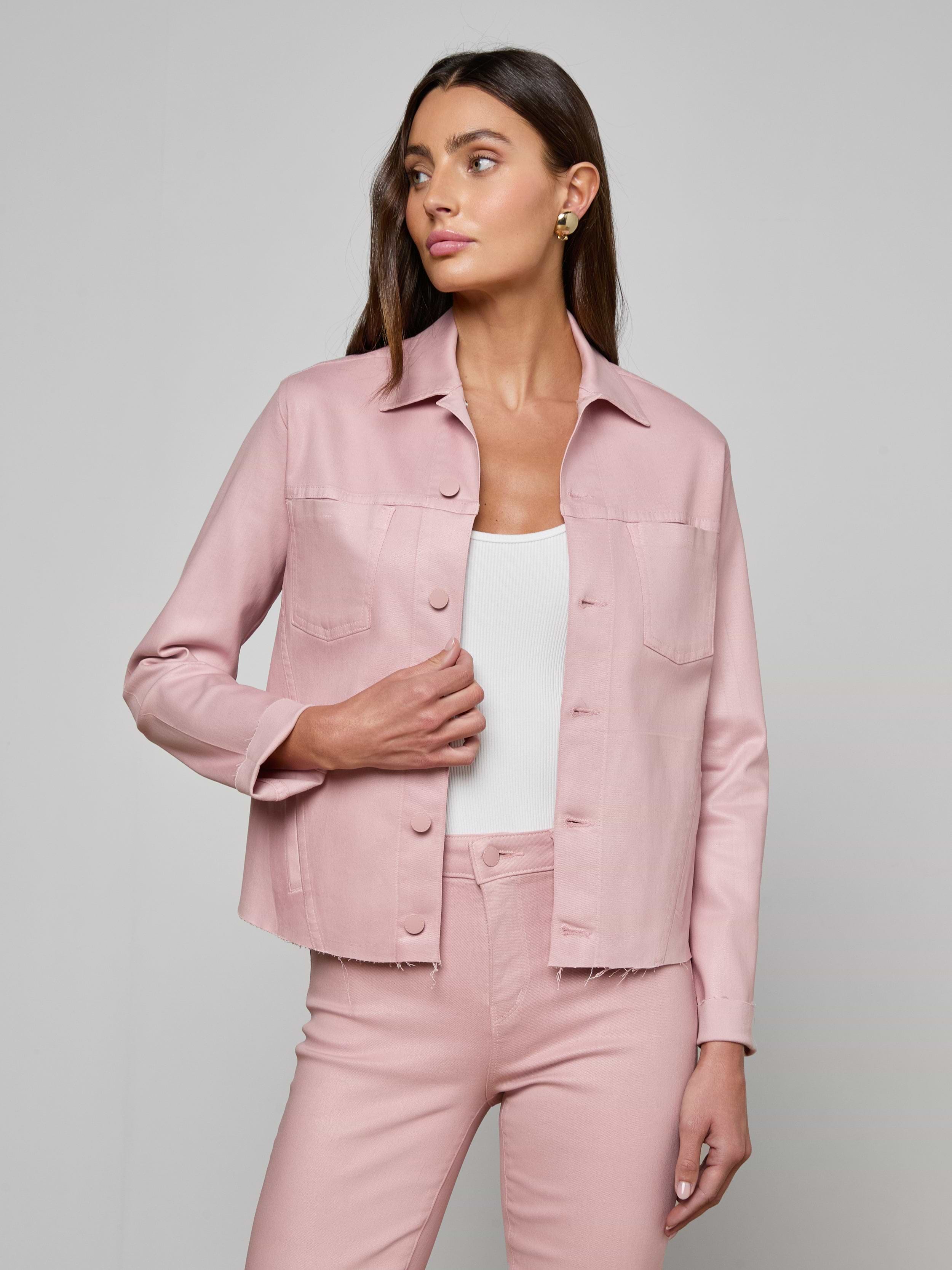 A woman wearing the Janelle Coated Jean Jacket in light pink with matching slim fit pants over a white top stands against a plain light background, looking to her left with one hand by her side and the other on her hip.