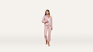 A woman wearing a light pink pantsuit and sandals stands on a plain white background, looking to her left with her hand on her jacket.