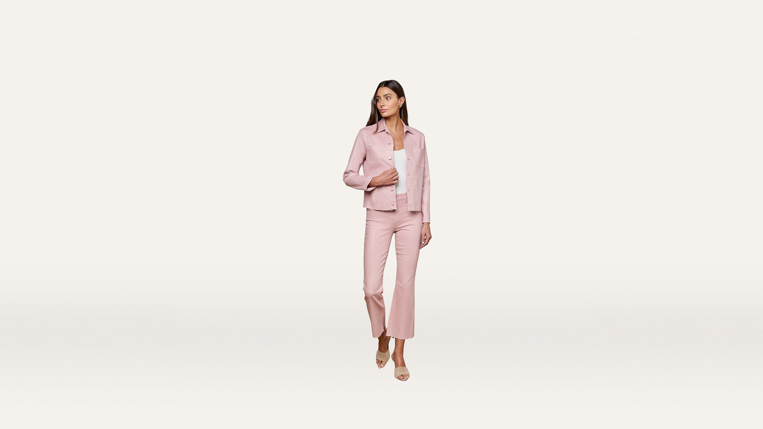 A woman wearing a light pink pantsuit and sandals stands on a plain white background, looking to her left with her hand on her jacket.