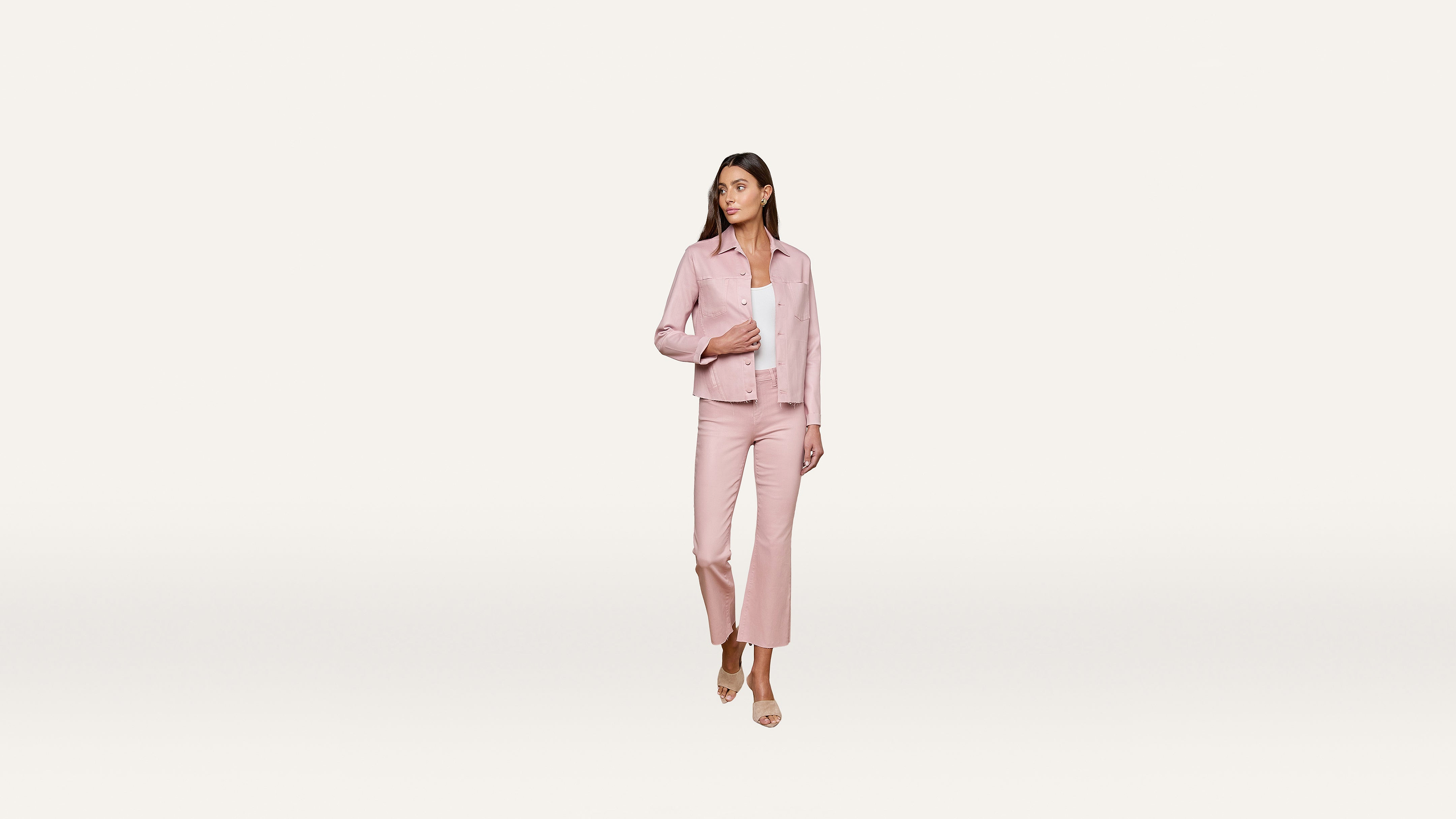 A woman wearing a light pink pantsuit and sandals stands on a plain white background, looking to her left with her hand on her jacket.