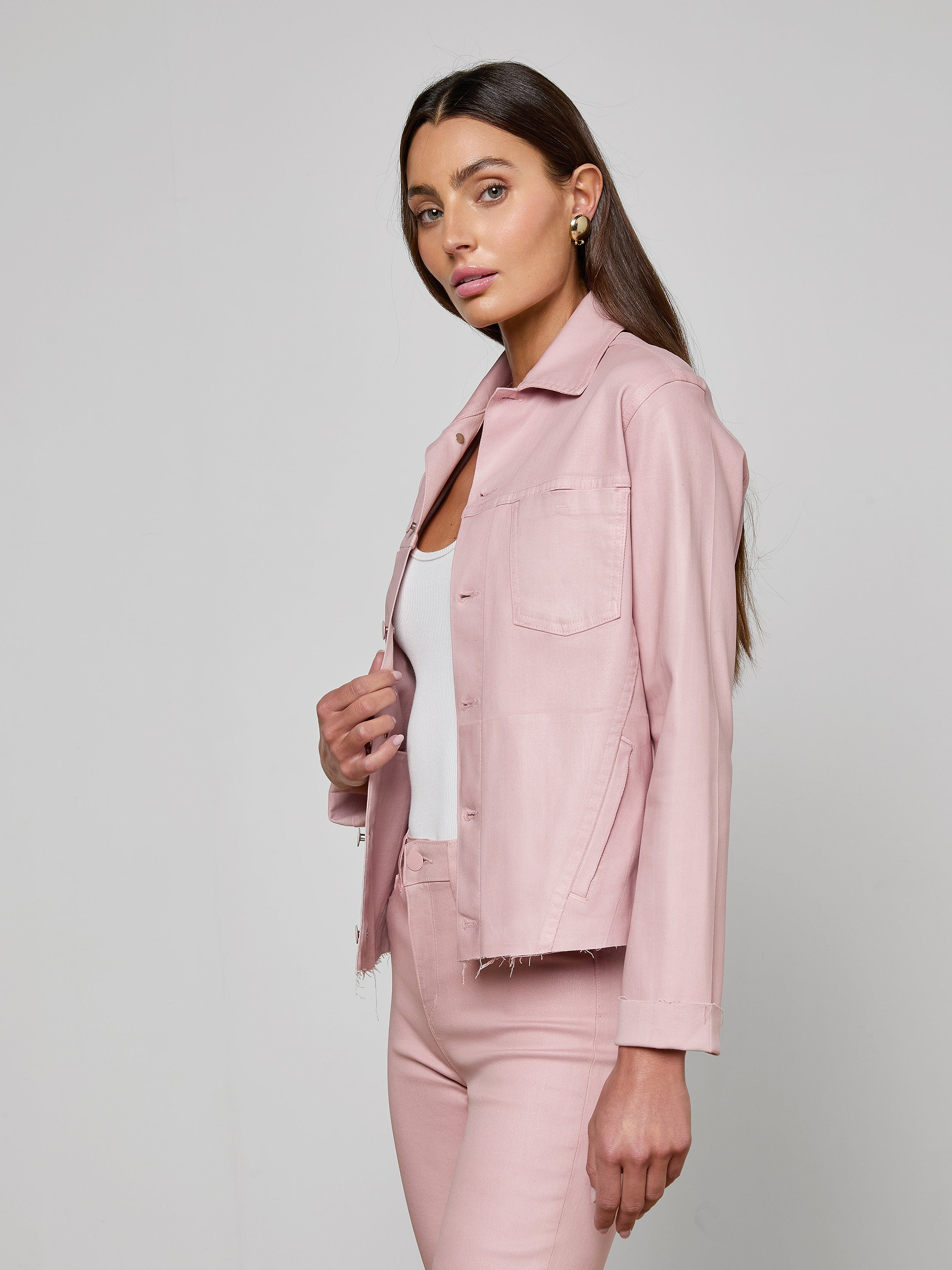 A woman with long brown hair wears the Janelle Coated Jean Jacket in light pink over a white top and matching pink pants, standing against a plain off-white background. She faces slightly to the side, looking forward with a neutral expression.