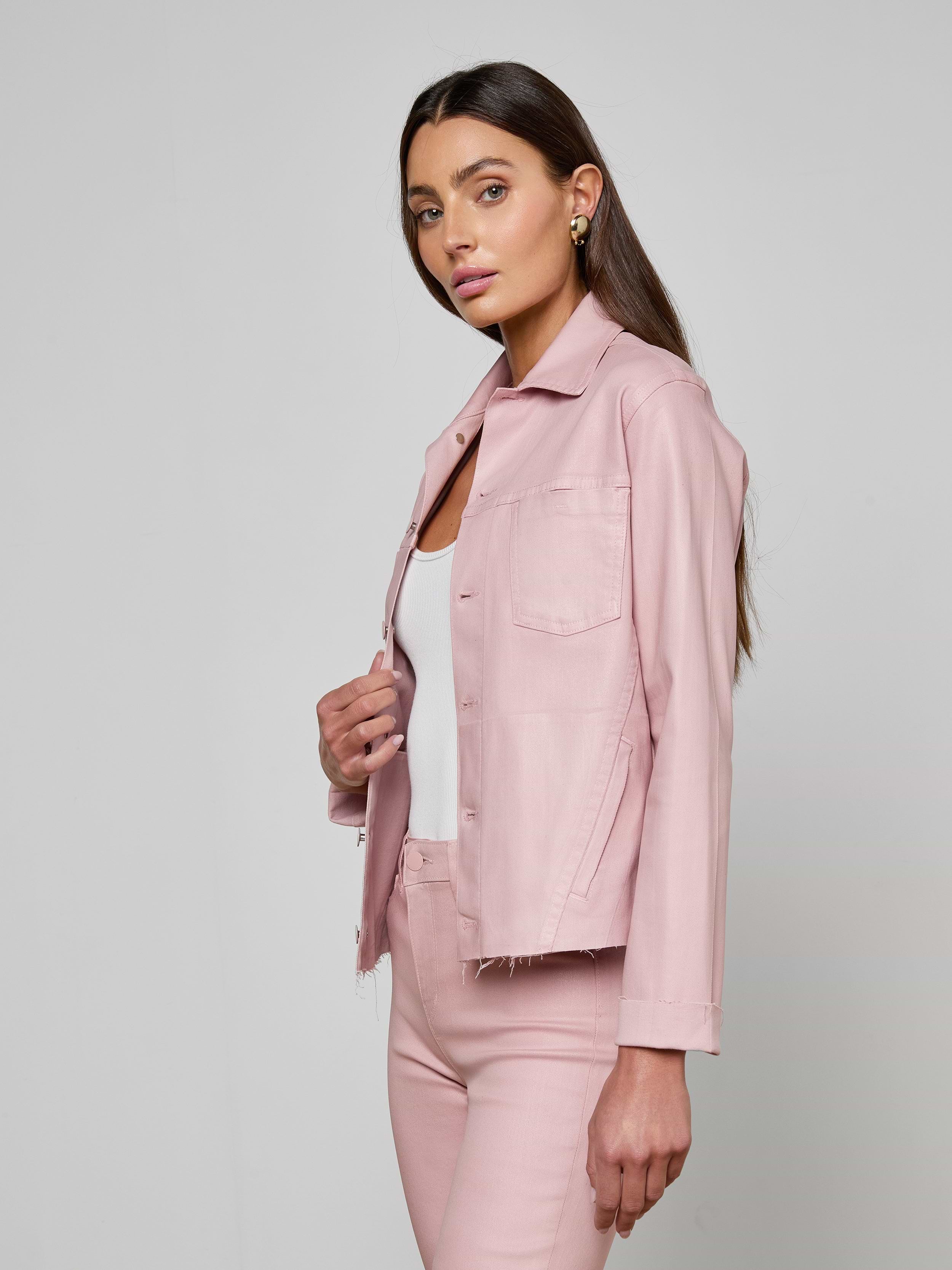 A woman with long brown hair wears the Janelle Coated Jean Jacket in light pink over a white top and matching pink pants, standing against a plain off-white background. She faces slightly to the side, looking forward with a neutral expression.