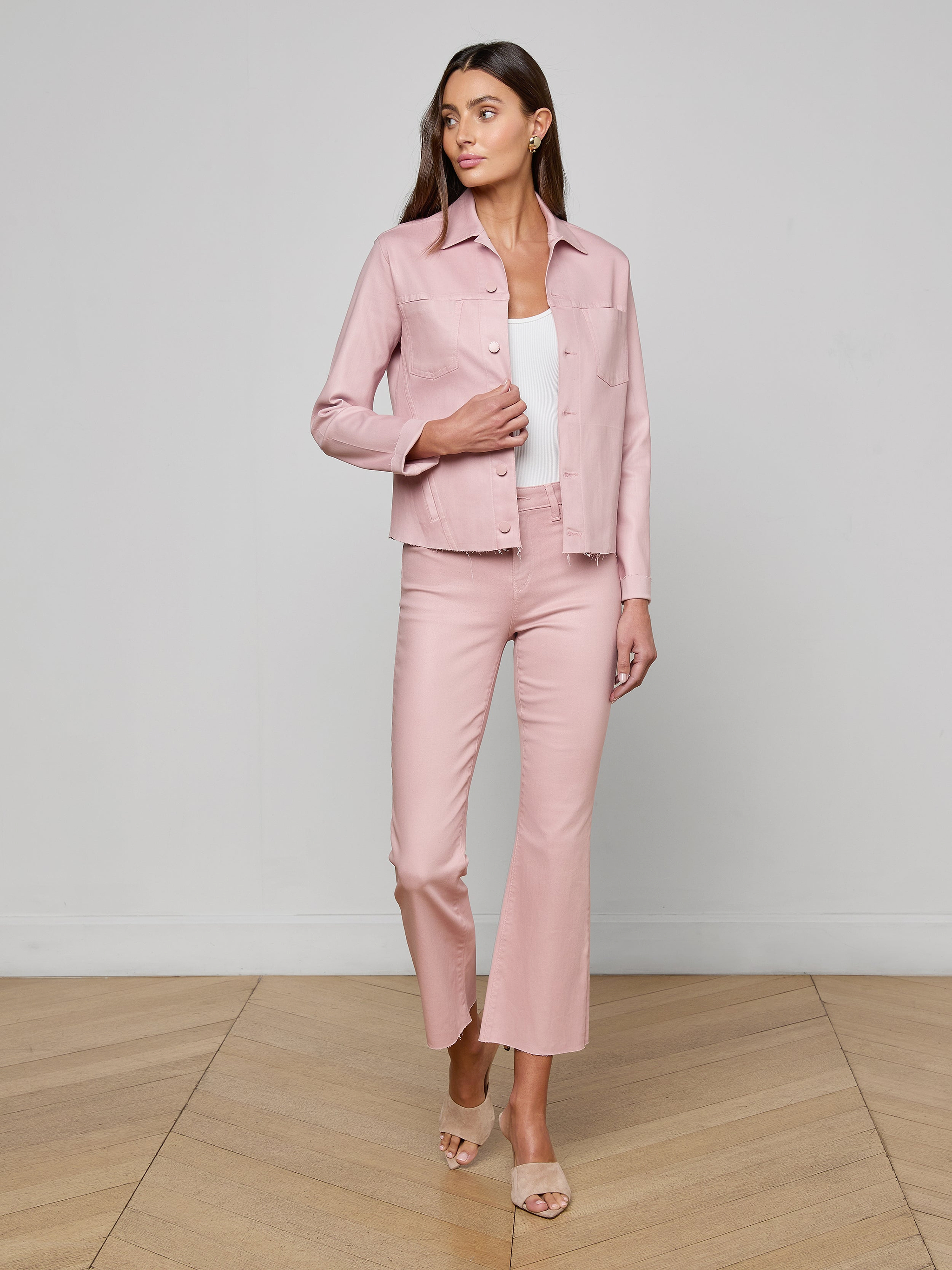 A woman stands against a plain white background, wearing the Janelle Coated Jean Jacket in light pink with matching pants over a white top. She has long hair and is wearing open-toed heels.