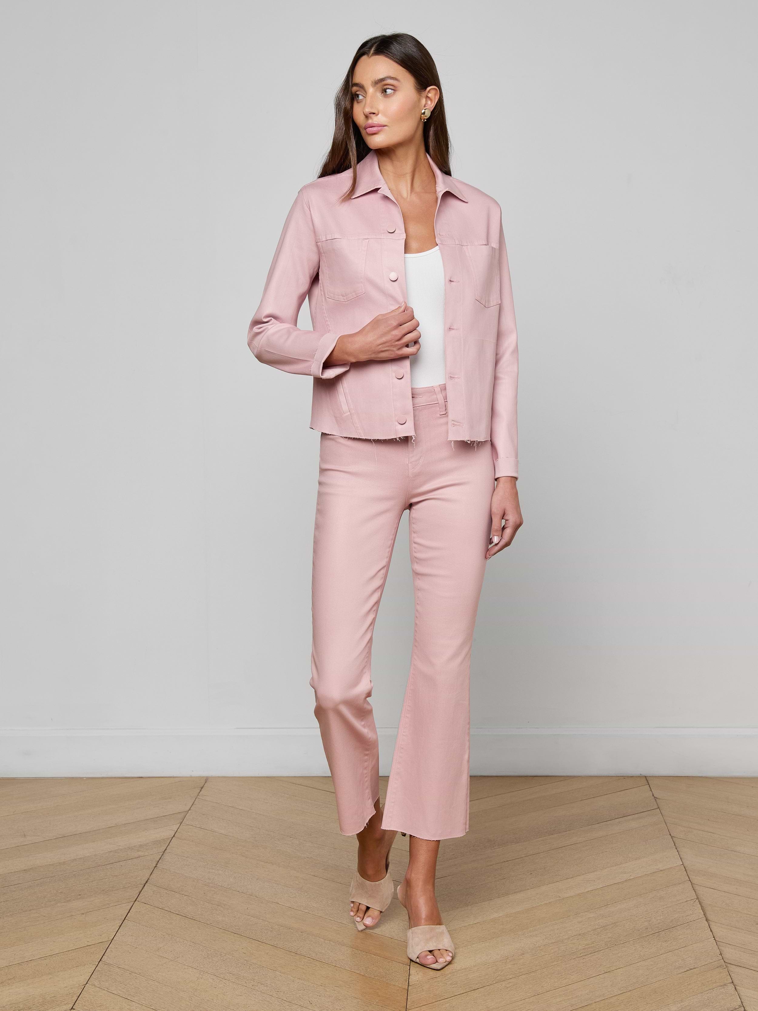 A woman stands against a plain white background, wearing the Janelle Coated Jean Jacket in light pink with matching pants over a white top. She has long hair and is wearing open-toed heels.