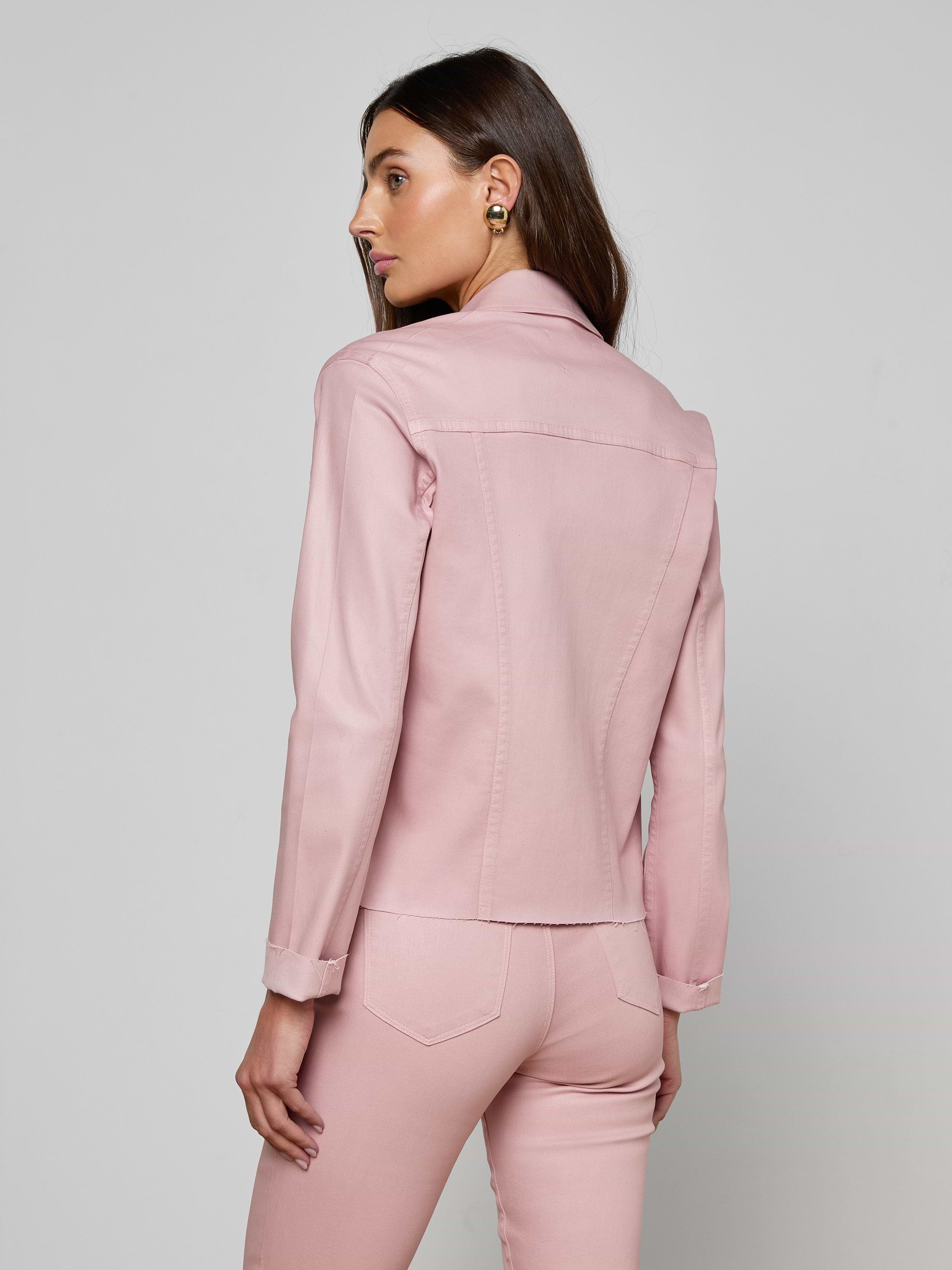 A woman with long brown hair wears the slim fit Janelle Coated Jean Jacket in light pink with matching pants, standing against a plain off-white background and looking to her left.