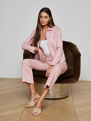 A woman with long brown hair sits on a brown chair wearing the Janelle Coated Jean Jacket over a white top and light pink pantsuit, paired with beige open-toe shoes. She gazes forward with a neutral expression in a minimal indoor setting.