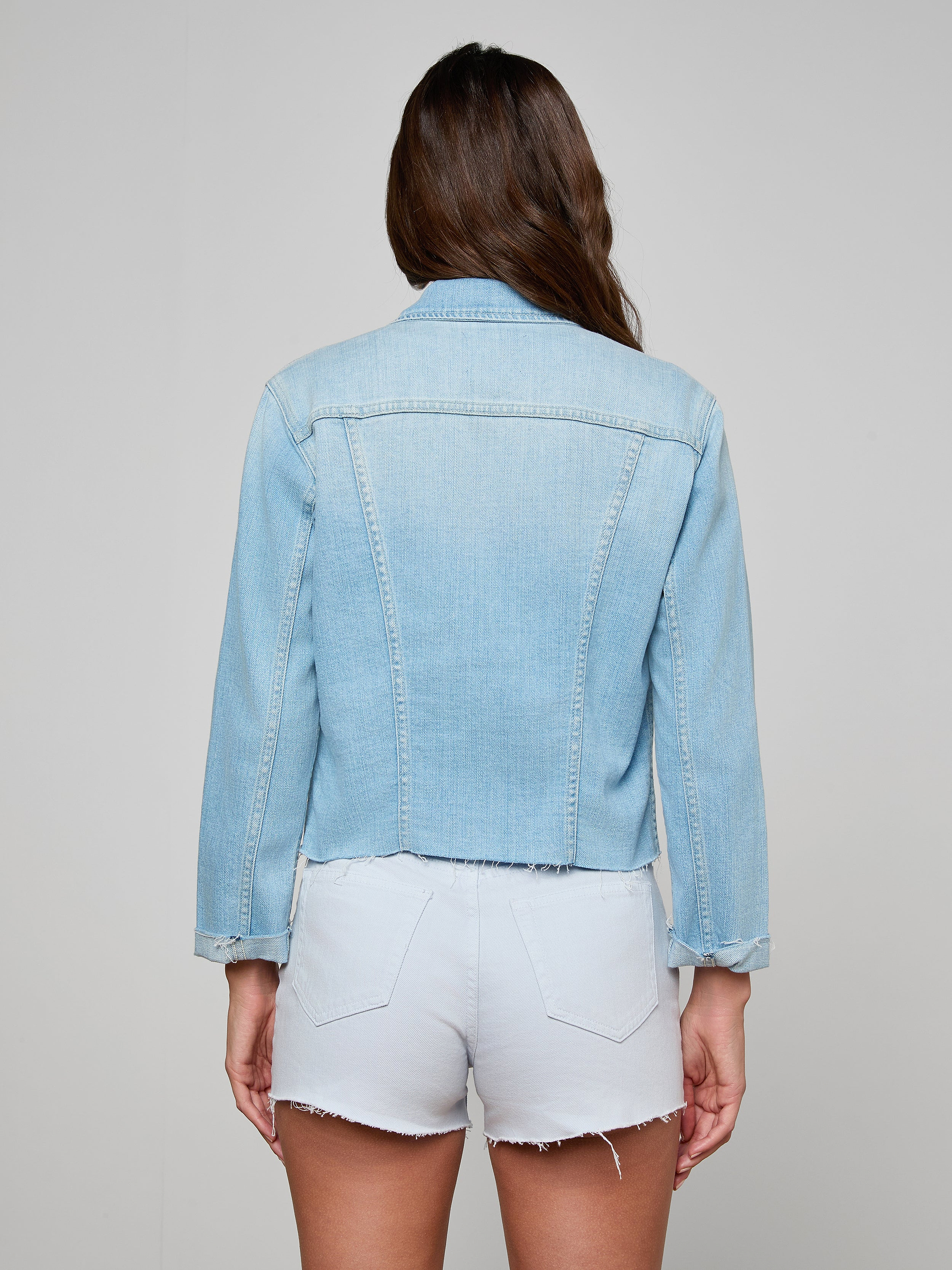 A woman with long brown hair, seen from behind, wears the Janelle Jean Jacket and white frayed hem shorts against a plain, light background.