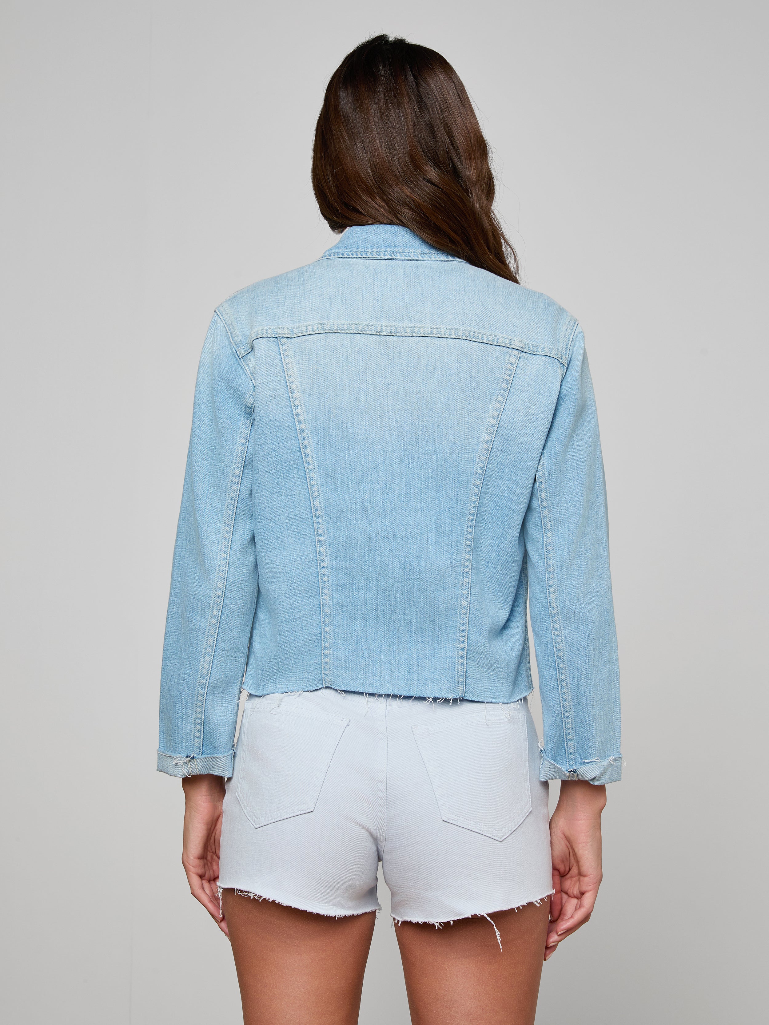 A woman with long brown hair, seen from behind, wears the Janelle Jean Jacket and white frayed hem shorts against a plain, light background.