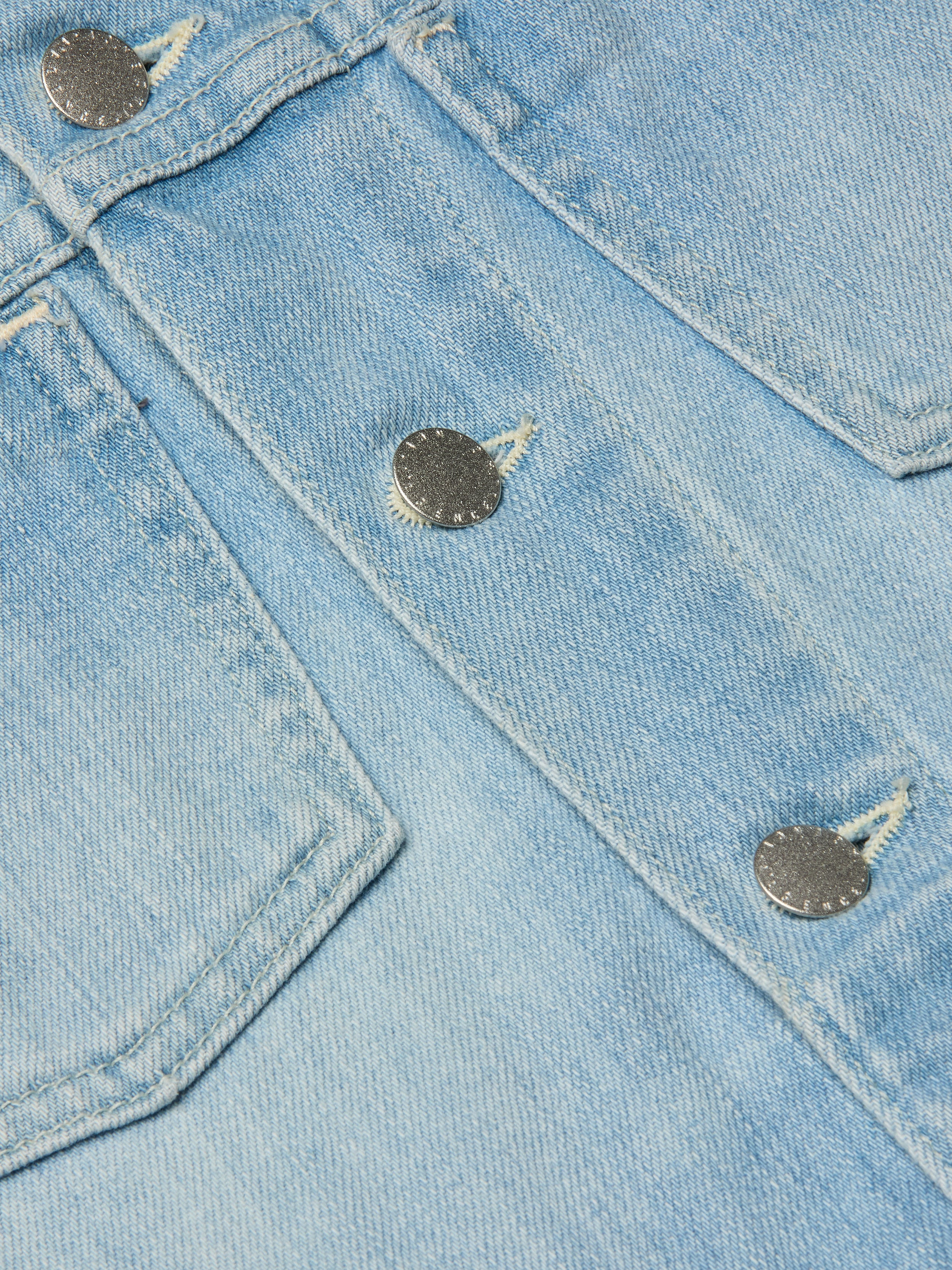 Close-up of light blue denim fabric with visible stitching and two metallic buttons, likely part of a pocket on the Janelle Jean Jacket.