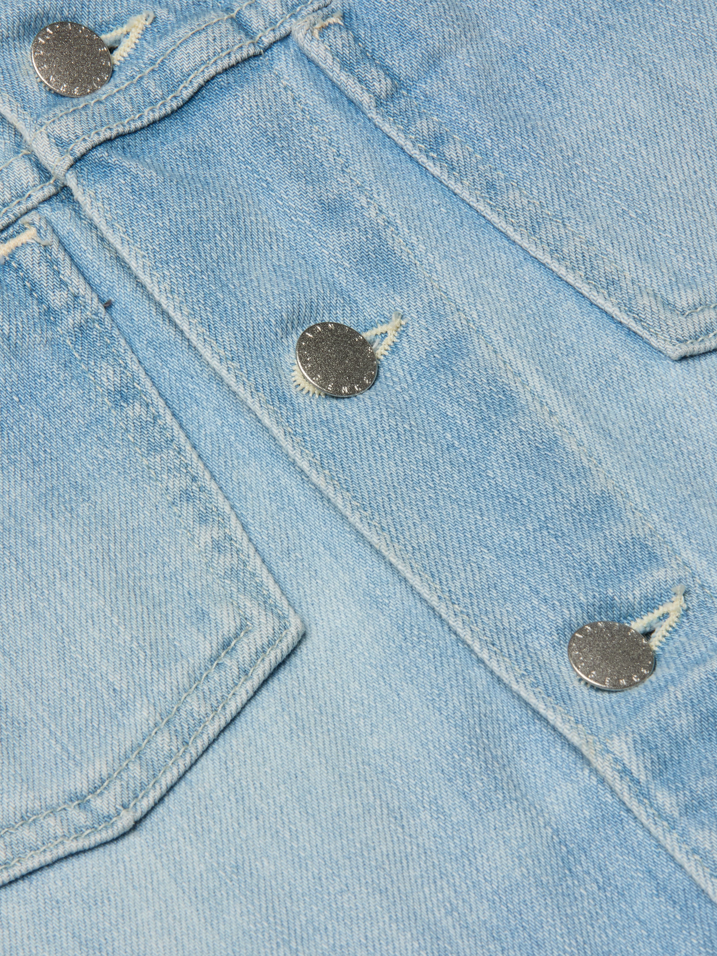 Close-up of light blue denim fabric with visible stitching and two metallic buttons, likely part of a pocket on the Janelle Jean Jacket.