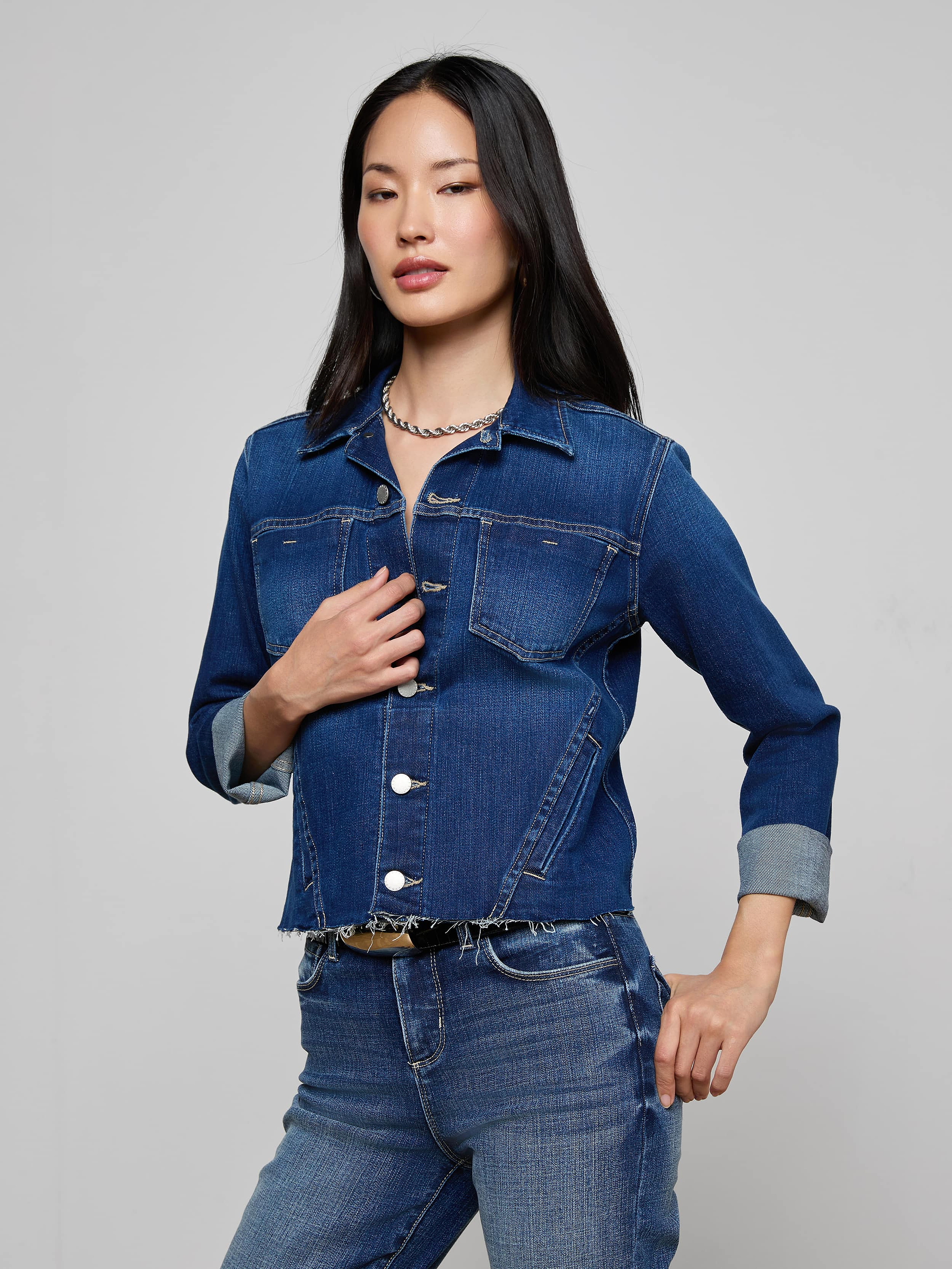 A woman wearing the Janelle Jean Jacket and matching indigo jeans poses with one hand on her waist and the other touching her jacket, set against a plain light background.