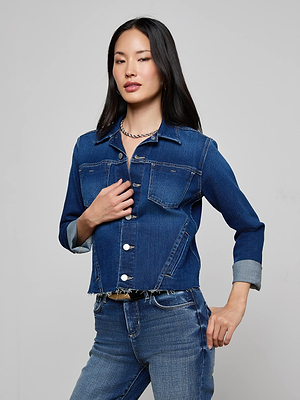 A woman with long black hair confidently wears the Janelle Jean Jacket, an indigo denim piece with 3/4 sleeves and a slim fit, paired with matching jeans. She stands against a light gray background, looking stylishly at the camera.