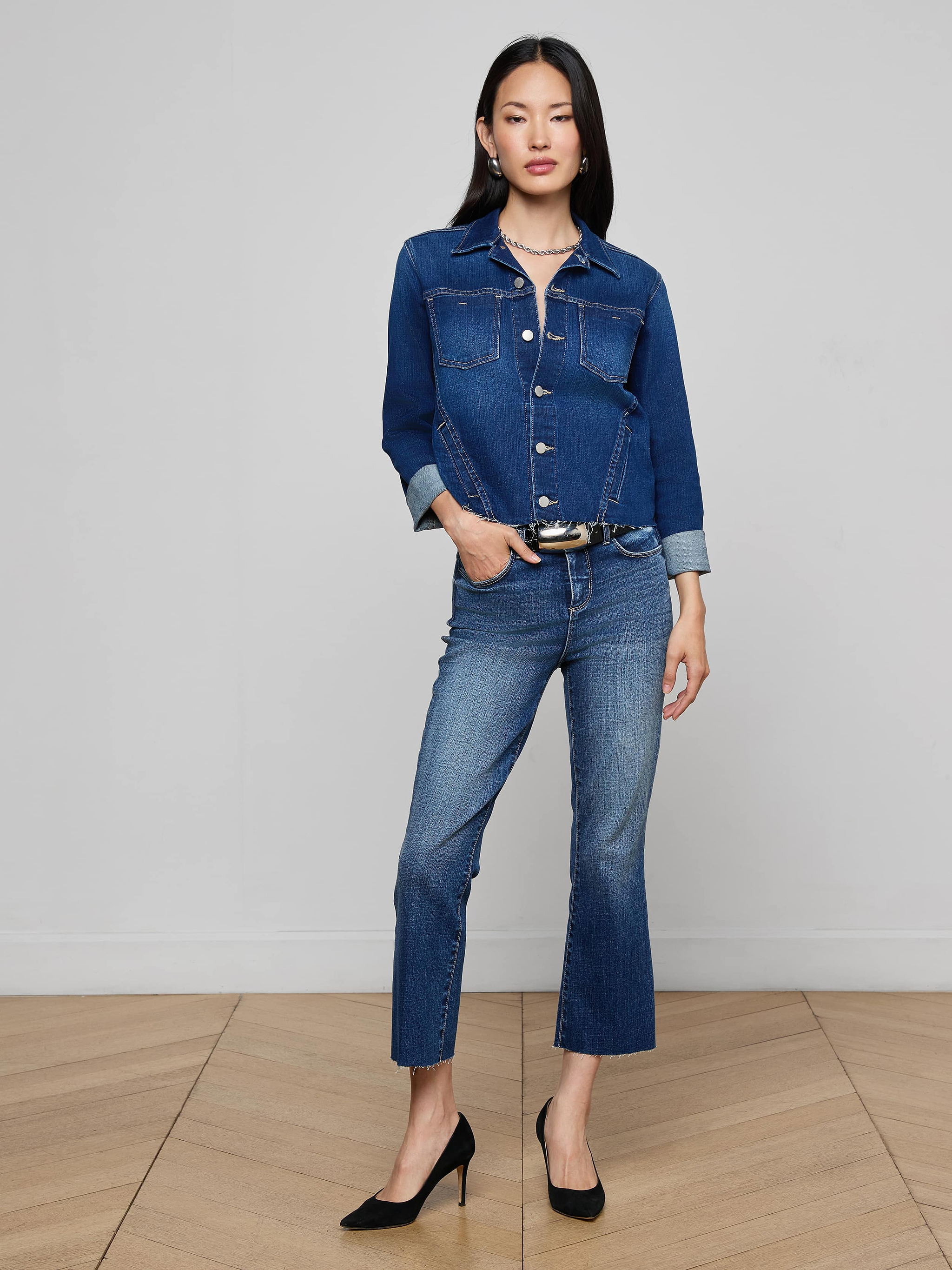 A woman stands against a plain background, wearing the Janelle Jean Jacket in indigo denim, matching jeans, and black heels, with one hand in her pocket.