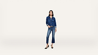 Woman standing against a plain white background, wearing a denim jacket and jeans with black high heels. She has one hand in her pocket and a neutral expression.
