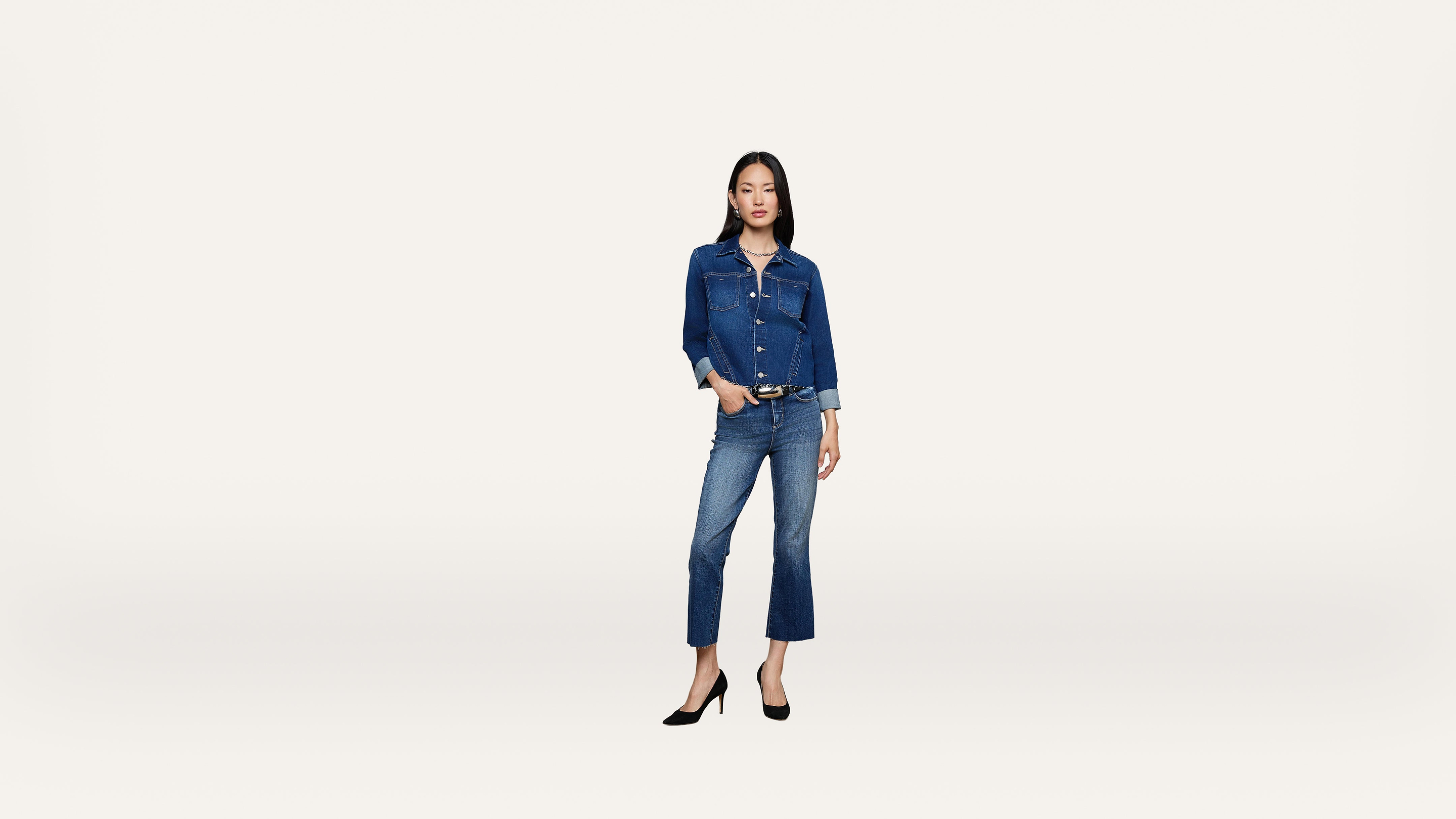 Woman standing against a plain white background, wearing a denim jacket and jeans with black high heels. She has one hand in her pocket and a neutral expression.