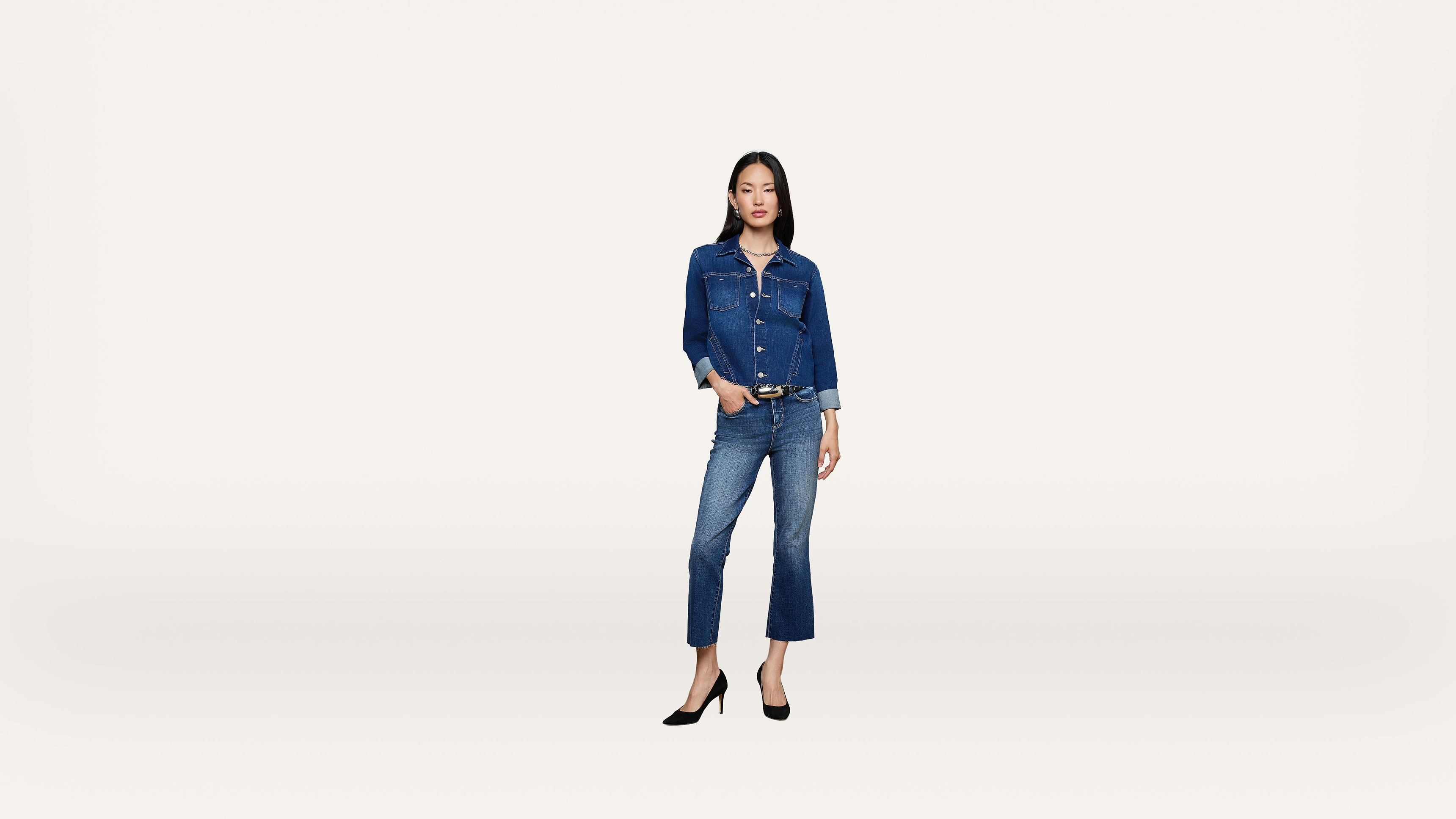 Woman standing against a plain white background, wearing a denim jacket and jeans with black high heels. She has one hand in her pocket and a neutral expression.