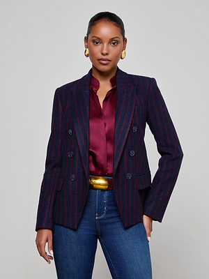 A woman stands against a plain background wearing the L'AGENCE Kenzie Pinstriped Blazer, a burgundy satin blouse, blue jeans, gold earrings, and a wide gold belt, looking confidently at the camera.