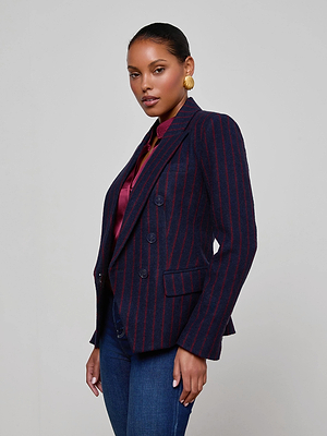 A woman with slicked-back hair models the L'AGENCE Kenzie Pinstriped Blazer—navy with red stripes—paired with a burgundy blouse, blue jeans, and gold hoop earrings against a plain light gray background.