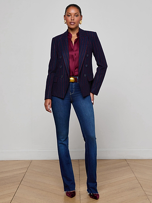 A woman stands against a plain wall wearing the Ruth Straight-Leg Jean in indigo with a raw hem, paired with a dark pinstripe blazer, burgundy blouse and heels, gold belt, and gold earrings. She stands on a wooden floor, looking confidently at the camera.