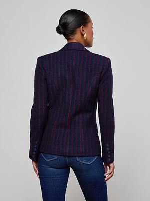 A woman faces away, wearing the L'AGENCE Kenzie Pinstriped Blazer with red stripes and dark blue jeans. Her hair is in a low bun, she sports gold hoop earrings, and stands against a plain light background.