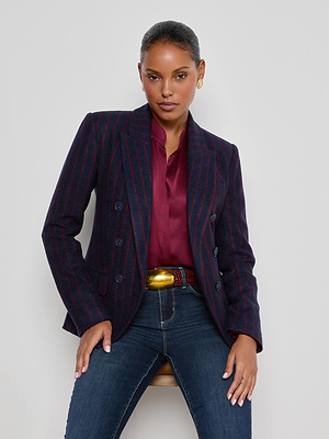 A woman in a maroon blouse, blue jeans, and a large gold belt buckle sits on a stool against a plain background, her L'AGENCE Kenzie Pinstriped Blazer adding a sharp, modern touch.