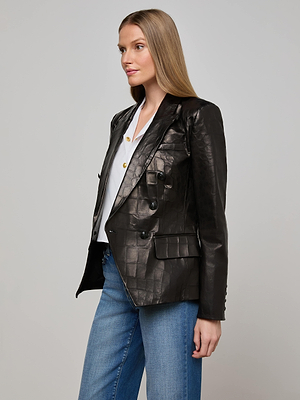 A woman with straight, light brown hair wears a black Kenzie Leather Blazer over a white shirt with gold buttons and blue jeans, standing against a plain light gray background.