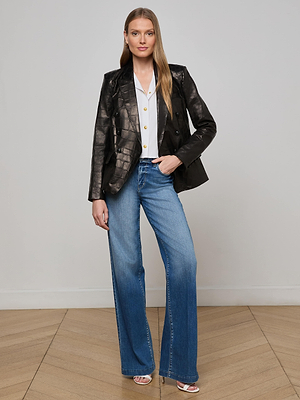 A woman stands indoors against a plain wall, wearing the Kenzie Leather Blazer in black croc-embossed leather, a white button-up blouse, blue wide-leg jeans, and white open-toe heels. She has long straight hair and a neutral expression.