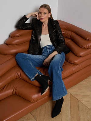 A woman sits on a modern brown leather couch, wearing the Kenzie Leather Blazer over a white top with blue flared jeans and black boots, casually crossing her legs and resting one arm on the back of the couch.