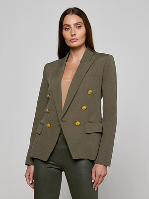 A woman with long brown hair wears the Kenzie Double-Breasted Blazer by L'AGENCE in olive, featuring gold buttons, over a beige top and matching olive pants, standing against a light gray background.