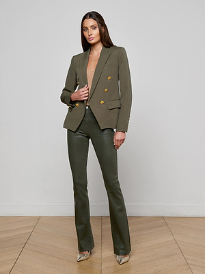 A woman poses on a wooden floor against a light gray wall wearing L'AGENCE's olive green Kenzie Double-Breasted Blazer with gold buttons, matching slim fit pants, a nude top, and silver heels.