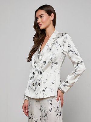 A woman with long brown hair models the Colin Silk Blazer in white charmeuse with a black abstract line print. She stands against a light gray background, gazing to the side with one hand on her hip.